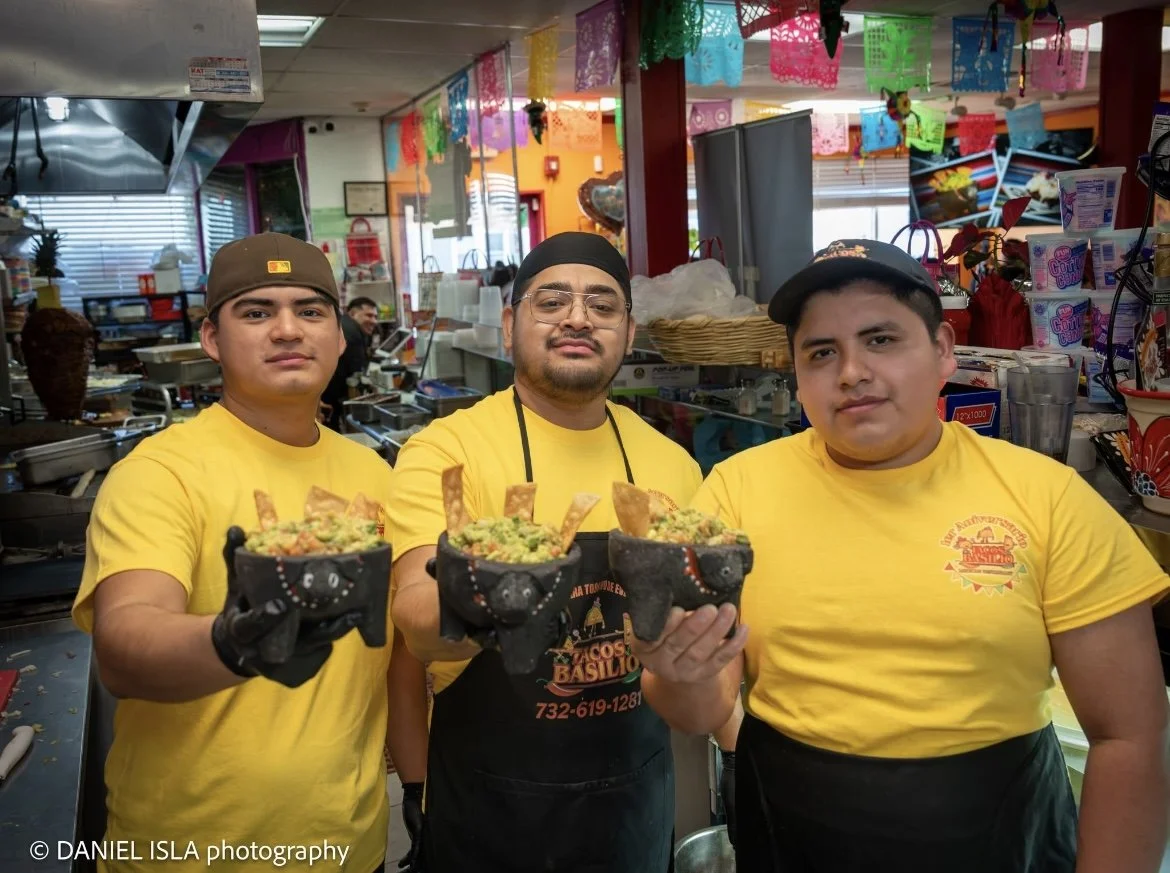 Three men wearing yellow T-shirts and black gloves holding homemade tacos with beef, guacamole, and chips in a colorful restaurant kitchen.
