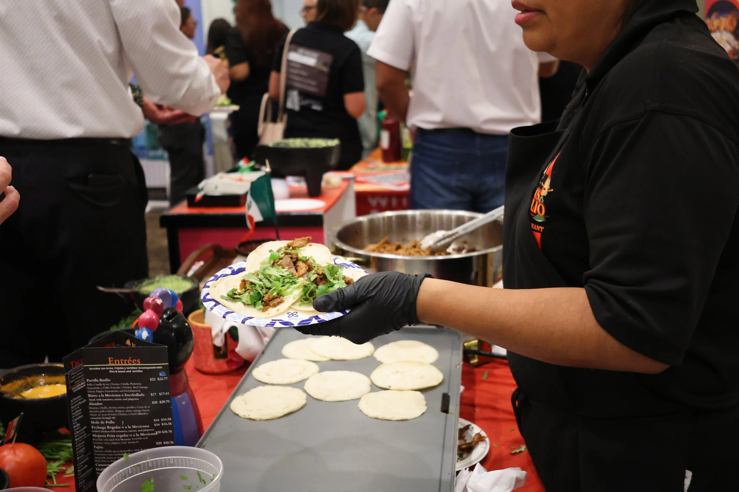 A person with black gloves serving food at a Mexican food event, with tacos filled with meat and garnished with cilantro on a tray, various ingredients, and condiments on the counter, and people in the background.