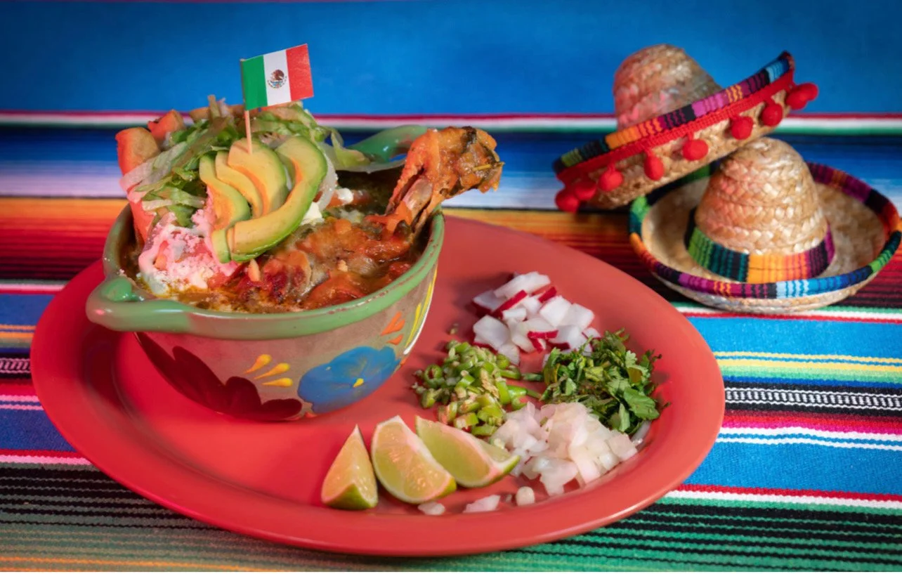 A colorful Mexican meal featuring a bowl of chicken tortilla soup garnished with avocado slices, a small Mexican flag, and a side of chopped radishes, onions, cilantro, and lime wedges on a red plate. Two small straw sombreros are in the background o