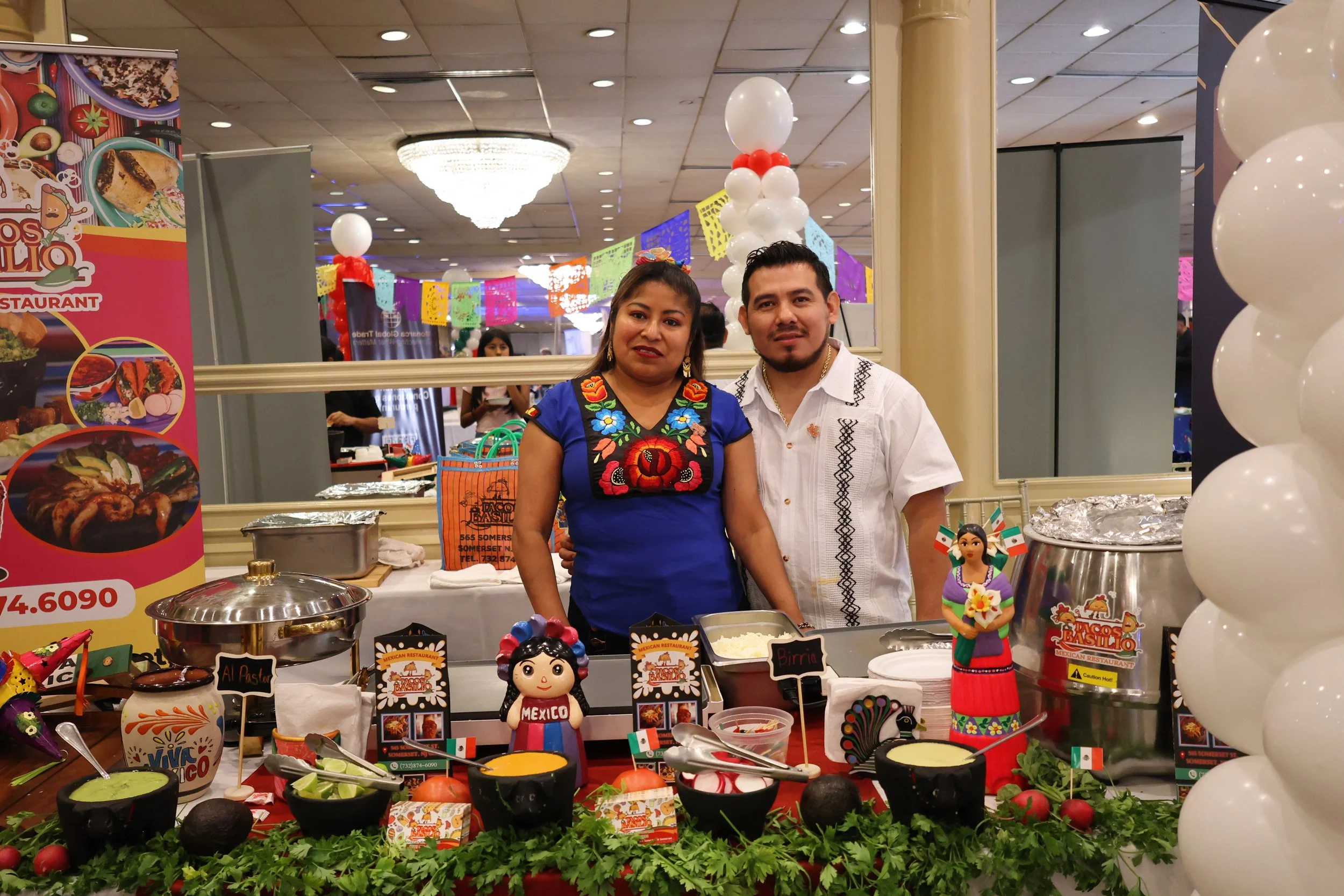 Two people, a woman and a man, stand behind a Mexican food stall decorated with colorful banners, balloons, and small Mexican flags. The woman wears a traditional embroidered blue dress, and the man wears a white shirt with black embroidery. The stall displays various Mexican dishes and figurines, including a doll labeled 'Mexico'. The background features a decorated banquet hall with balloons and a chandelier.
