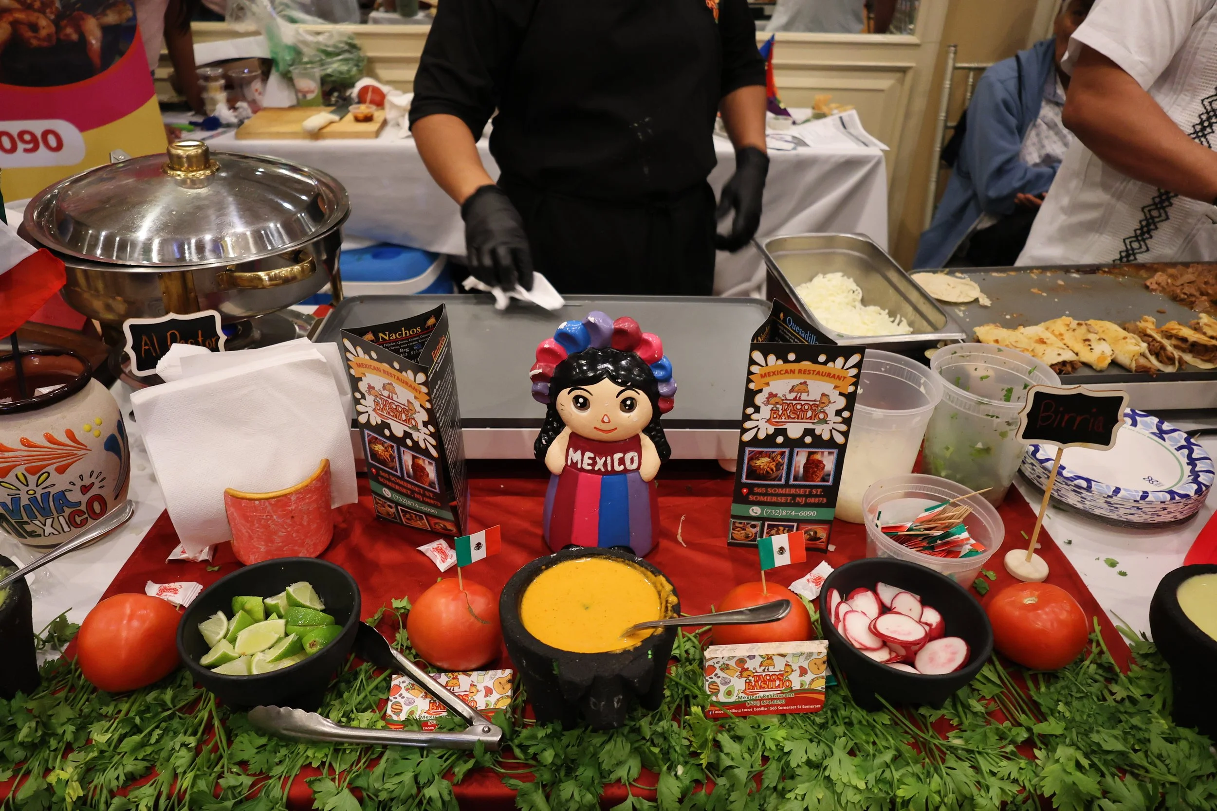 Display of Mexican food ingredients and dishes on a table, including chopped vegetables, radishes, tomatoes, lime wedges, and bowls of sauces, with decorative elements such as a small Mexican figurine and flags.