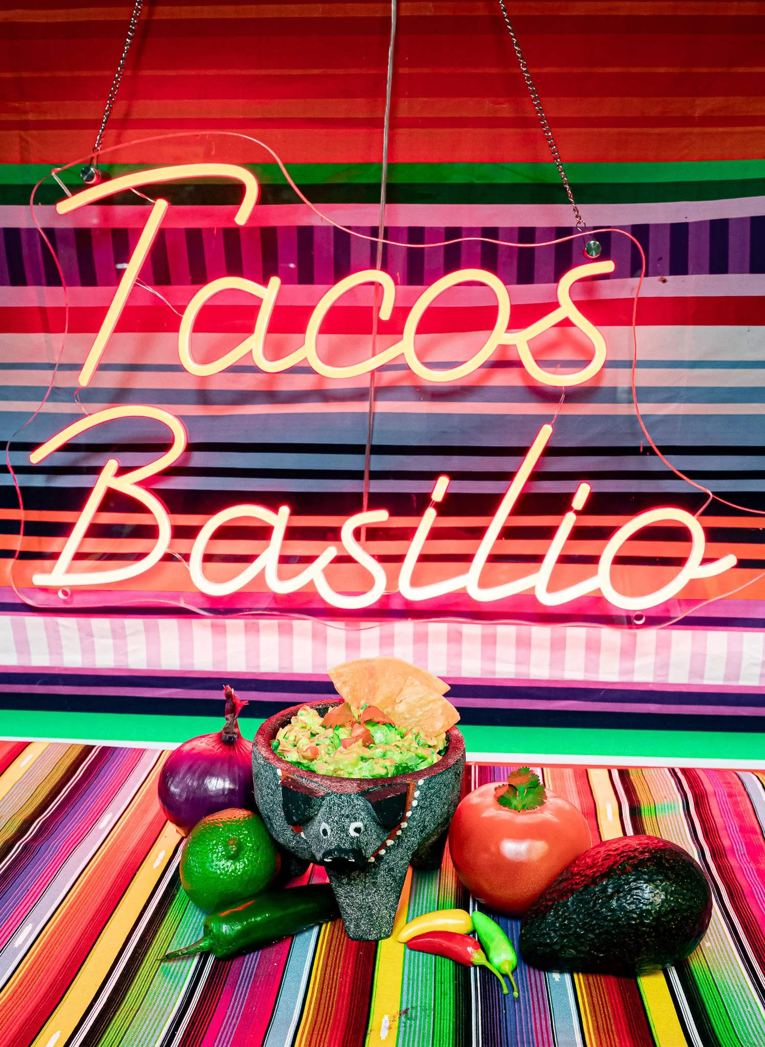Colorful table display with a neon sign that says "Tacos Bachi," a bowl of guacamole with a tortilla chip, and several avocados, limes, and chili peppers.
