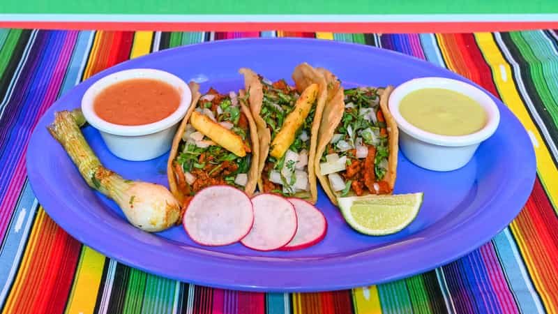 Three tacos with shredded lettuce, cheese, and meat on a blue plate, served with radish slices, lime wedges, and two small bowls of sauce, on a colorful striped tablecloth.