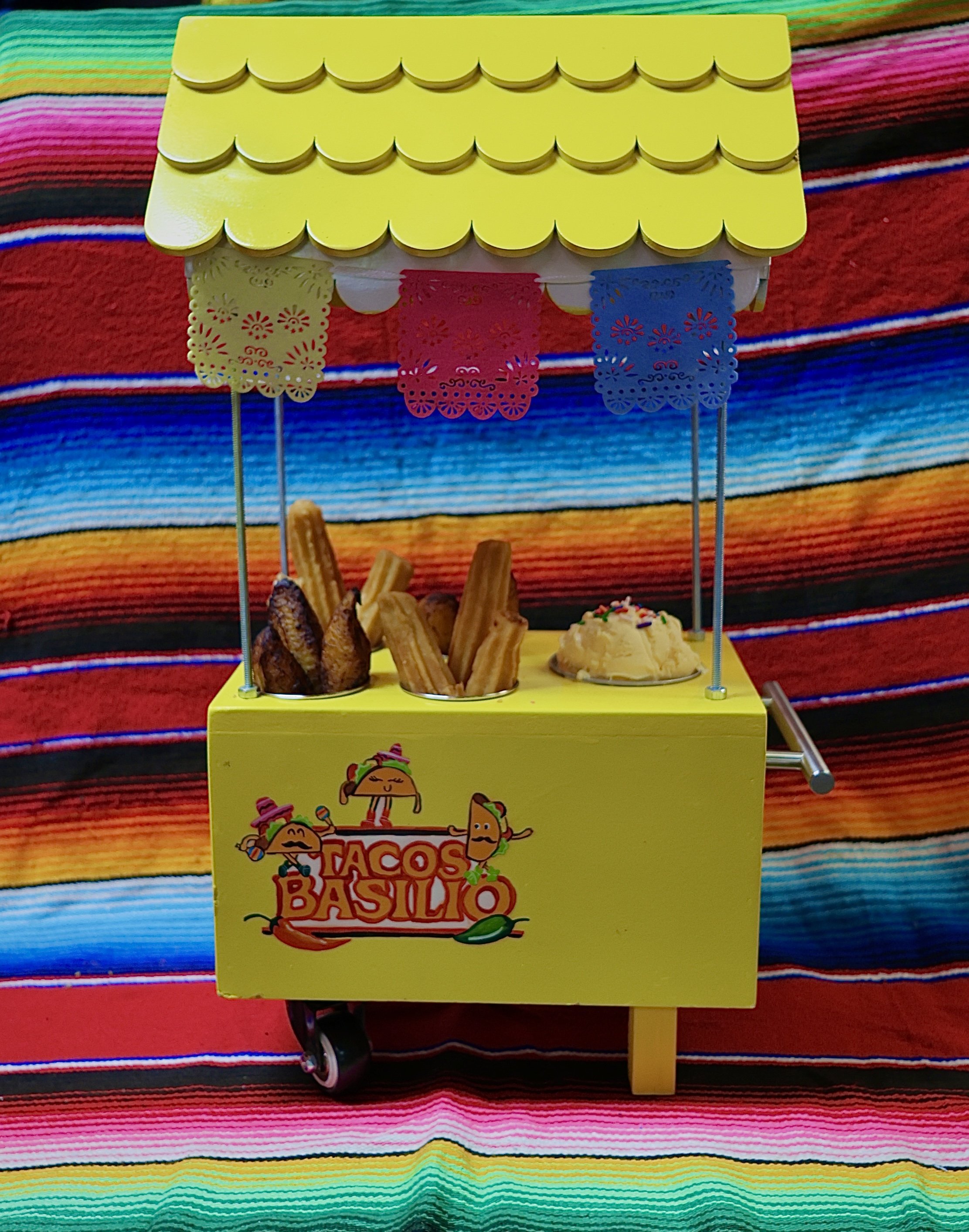 A yellow decorative box with colorful papel picado banners hanging from its roof, featuring a taco character with a smiley face. The box has the words "Tacos Basillo" with images of tacos and various festive decorations on its sides. It is placed on 