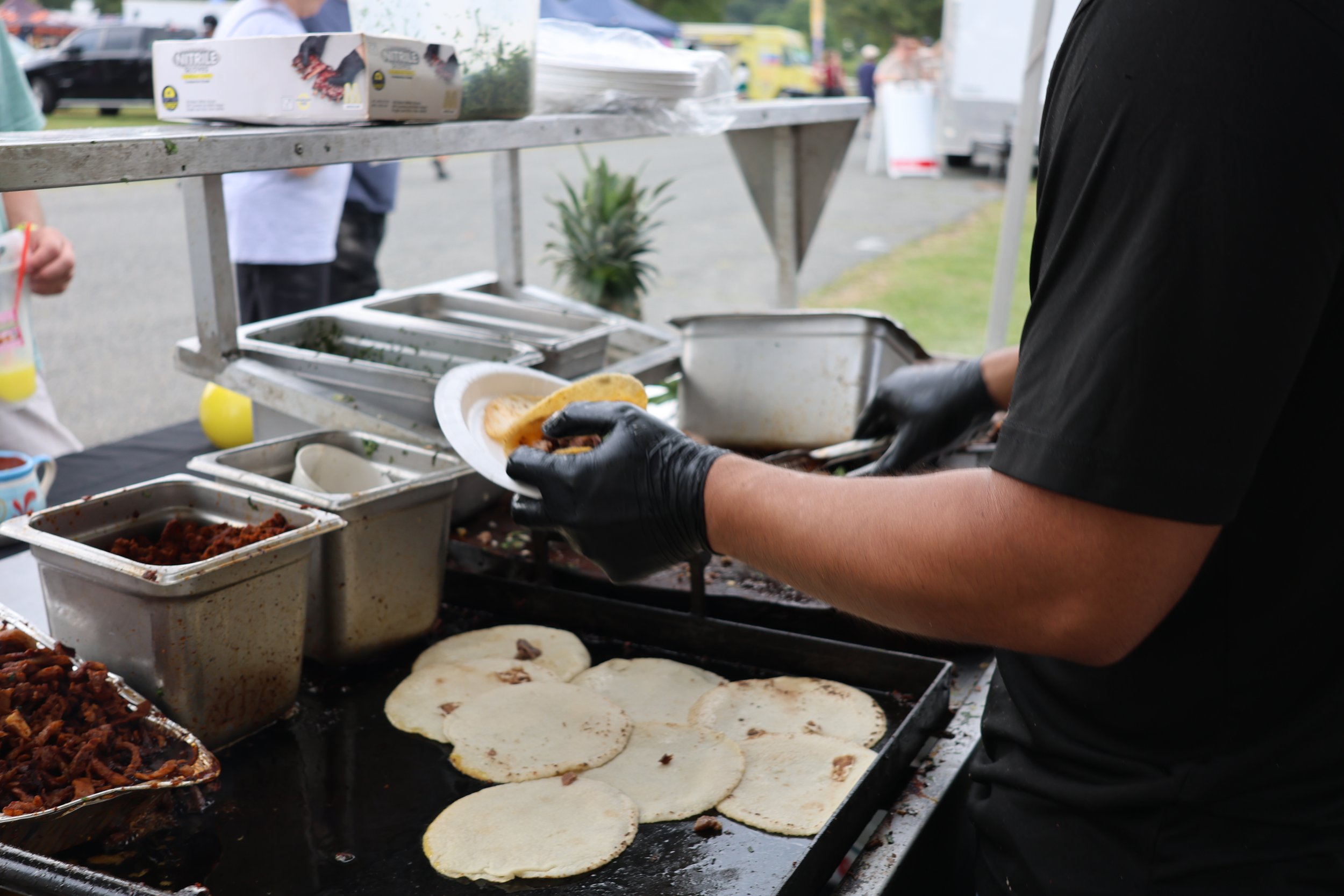 A person wearing black gloves preparing tacos with tortillas, meat, and toppings at an outdoor food stall.