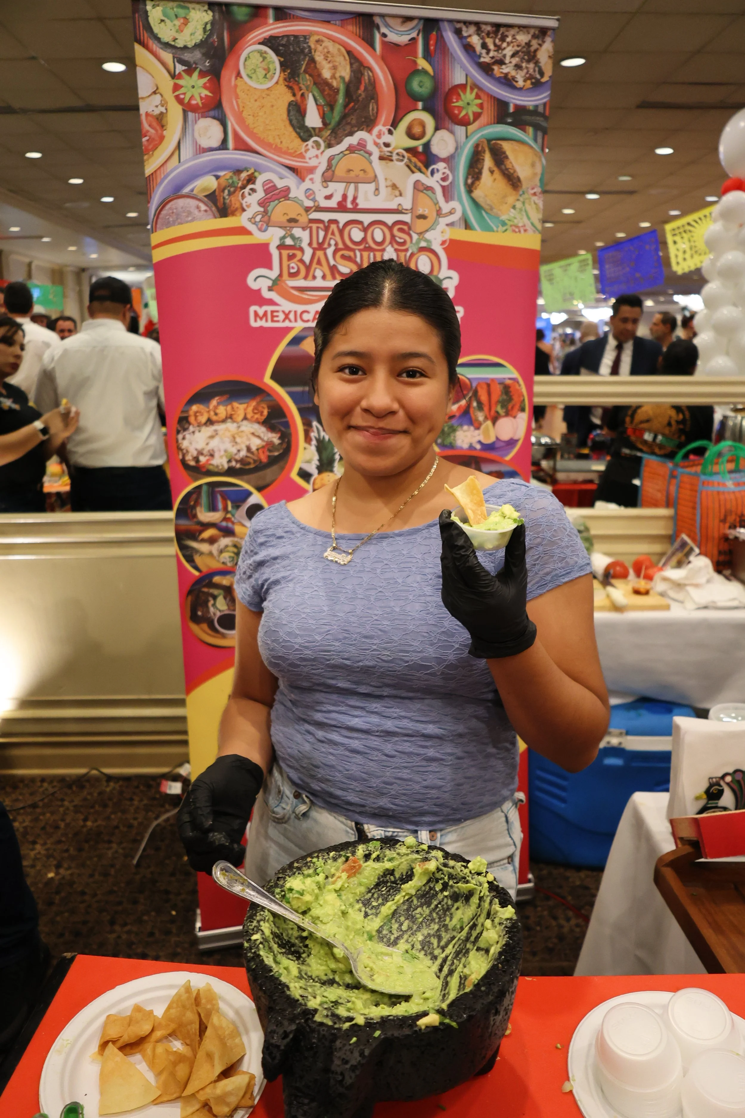 A woman smiling at a food stall holding a small bowl of guacamole. She is standing in front of a large bowl of freshly made guacamole with a spoon. Behind her is a colorful banner with images of Mexican dishes and a crowd of people at the event.