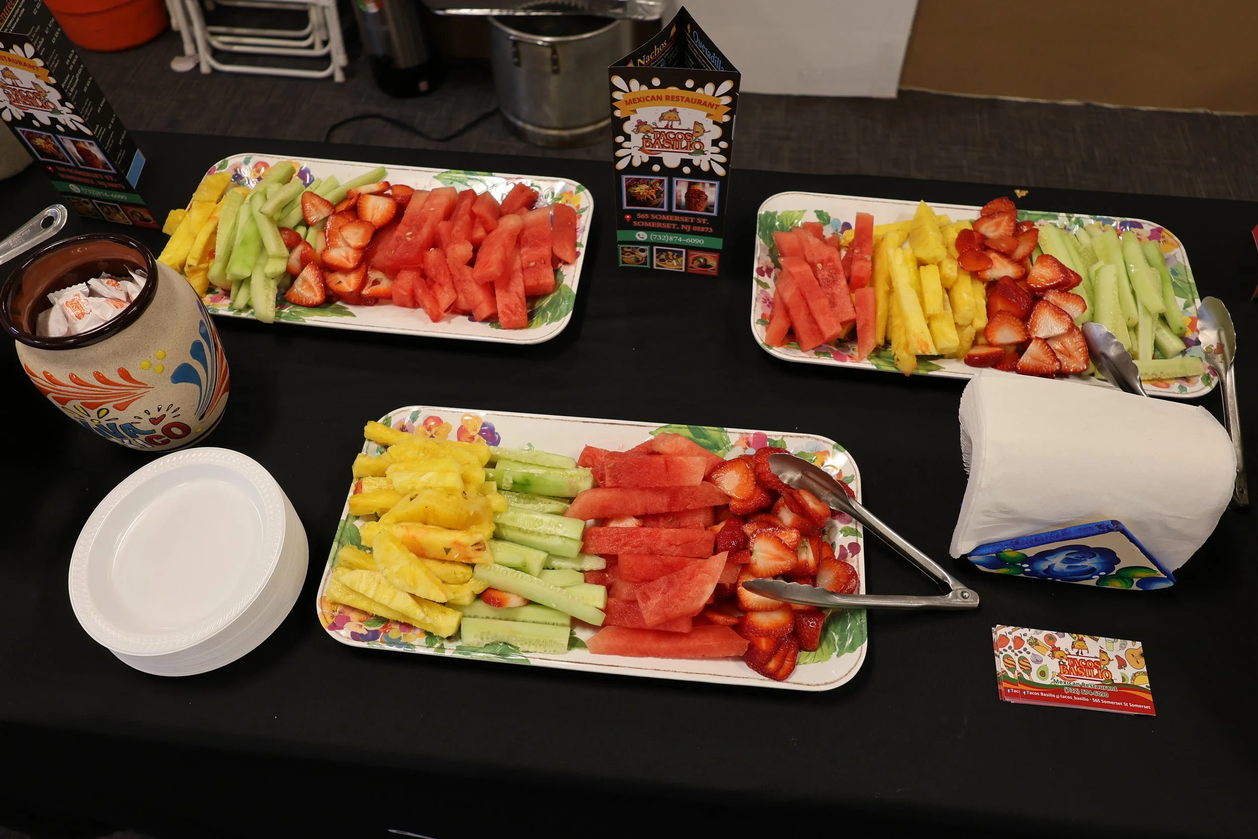Assorted sliced fresh fruits on decorative trays, including watermelon, strawberries, pineapple, yellow melon, and honeydew, on a black table with a napkin holder, paper plates, and promotional cards for a Mexican restaurant.