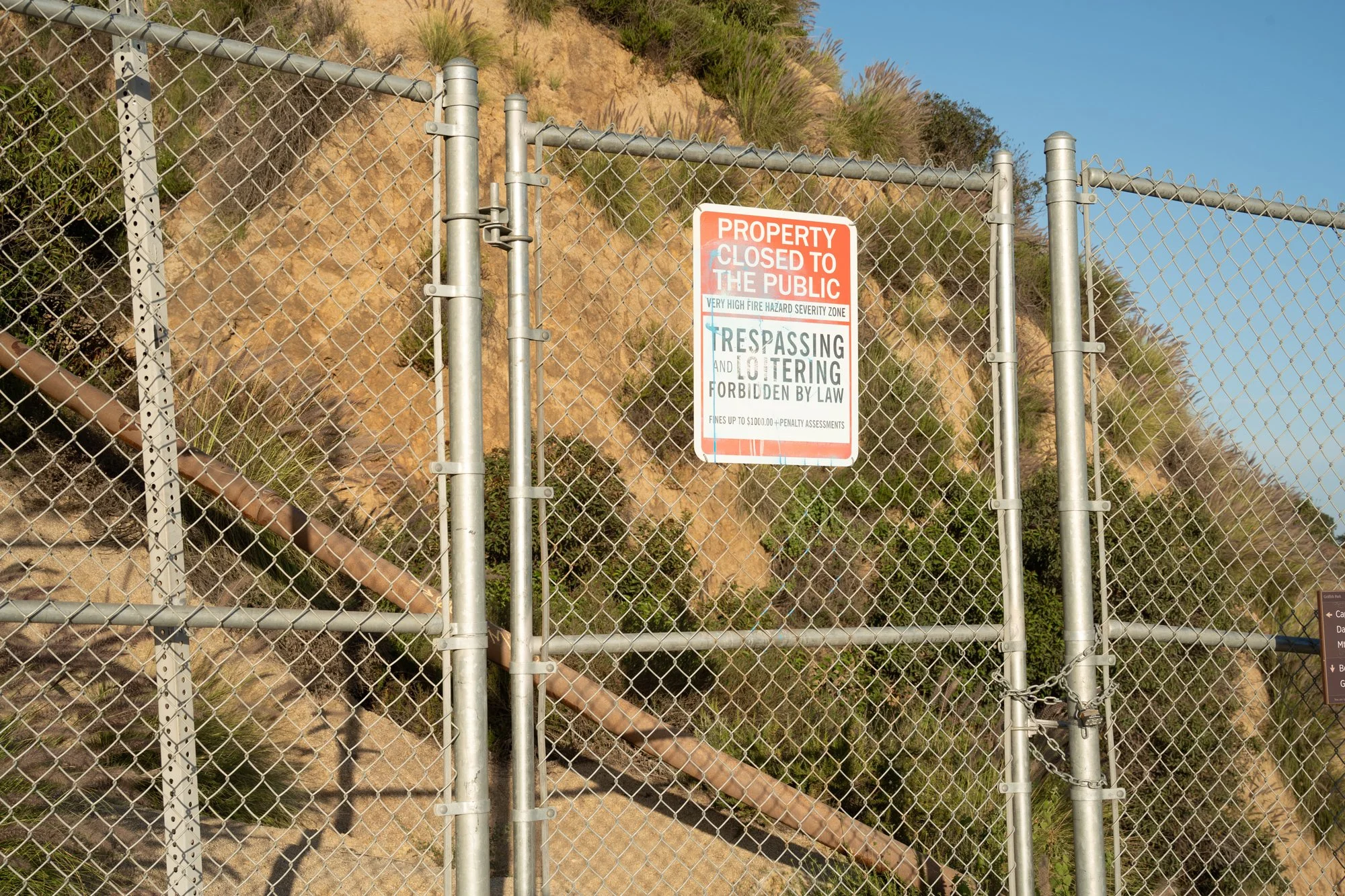 A metal chain-link fence with a sign that reads 'Property closed to the public. Trespassing and loitering forbidden by law.' Behind the fence, there is a hillside with rocks and sparse vegetation, and a clear blue sky above.