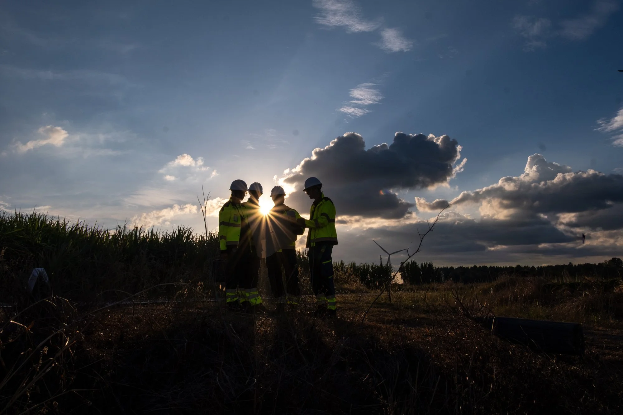 Four construction workers wearing safety helmets and vests standing in a field at sunset, silhouetted against the sky with clouds.