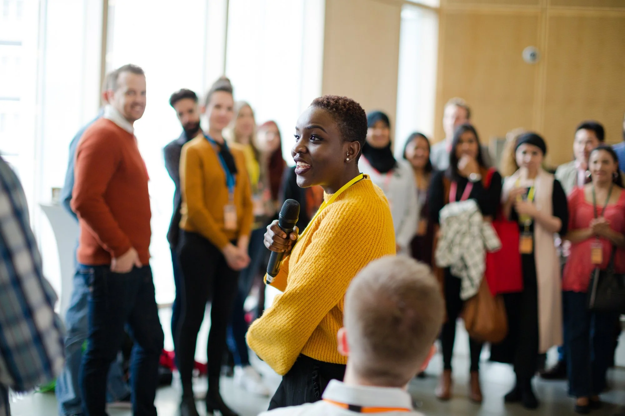 A woman in a yellow sweater holding a microphone speaking to an audience at a conference or event, with a diverse group of people listening.
