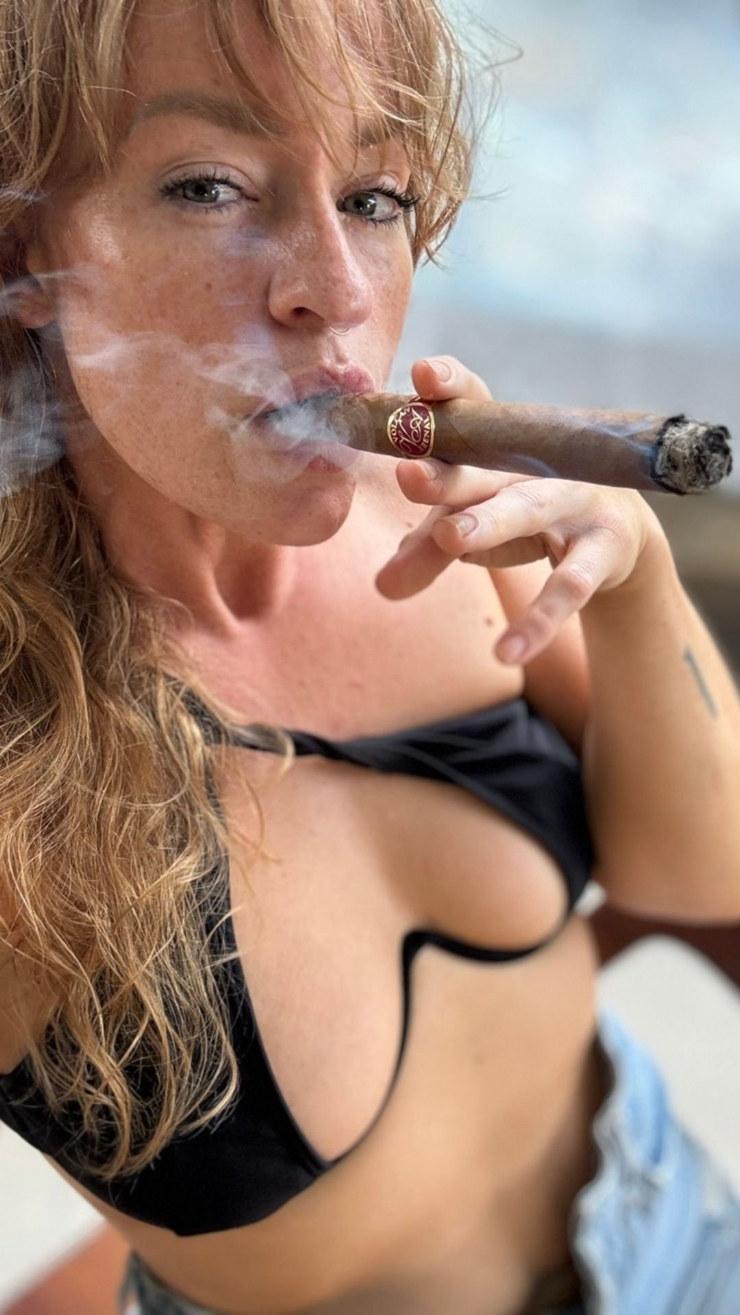 Woman with freckles and wavy hair smoking a cigar, wearing a black top with a deep cut and jeans.