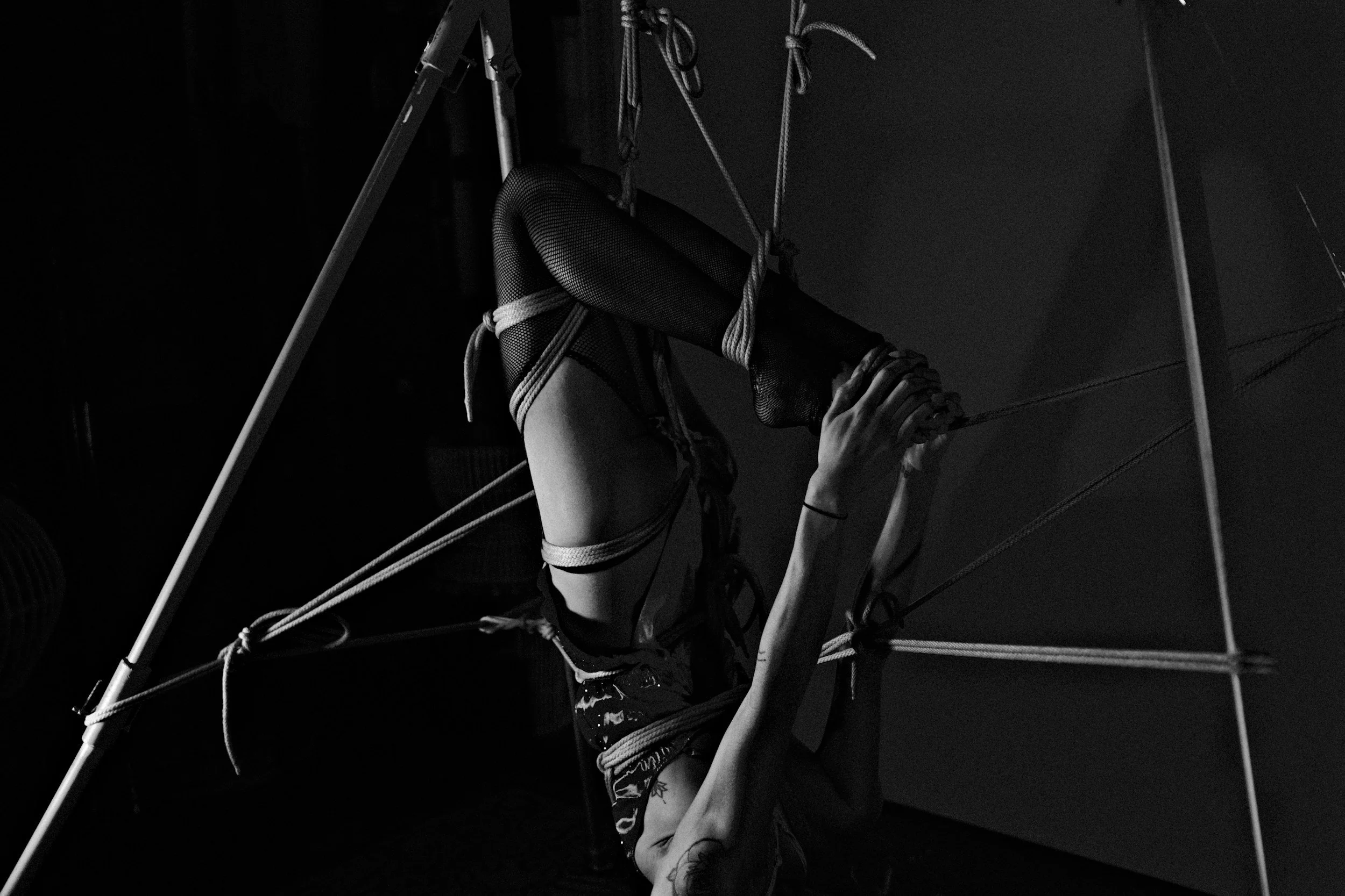 A person performing an aerial acrobatic pose on a trapeze, hanging upside down with legs hooked around the trapeze and hands holding the ropes, in a dark setting.