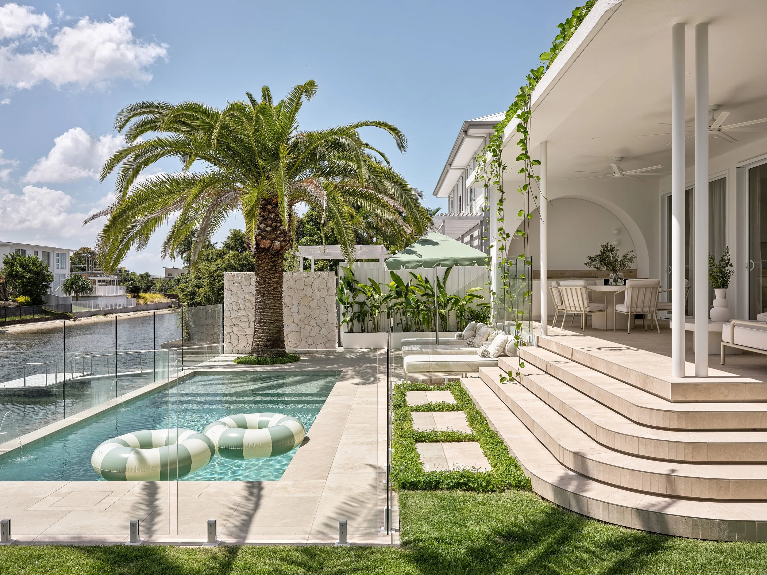 Luxury backyard with swimming pool, palm tree, outdoor furniture, and a waterway with modern houses in the background.