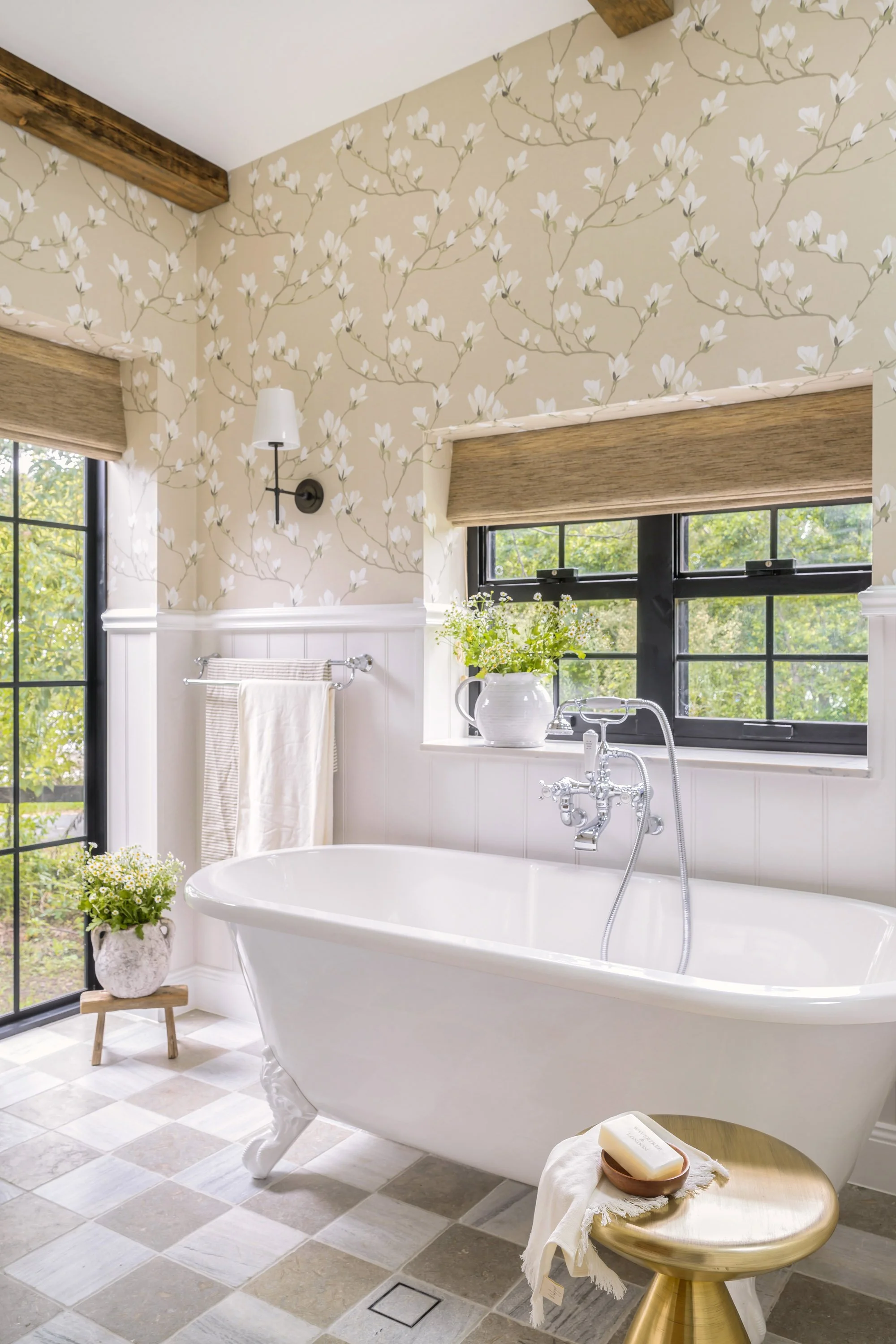 A bright bathroom with a clawfoot bathtub, a window with a greenery view, and floral wallpaper with white flowers and beige background.