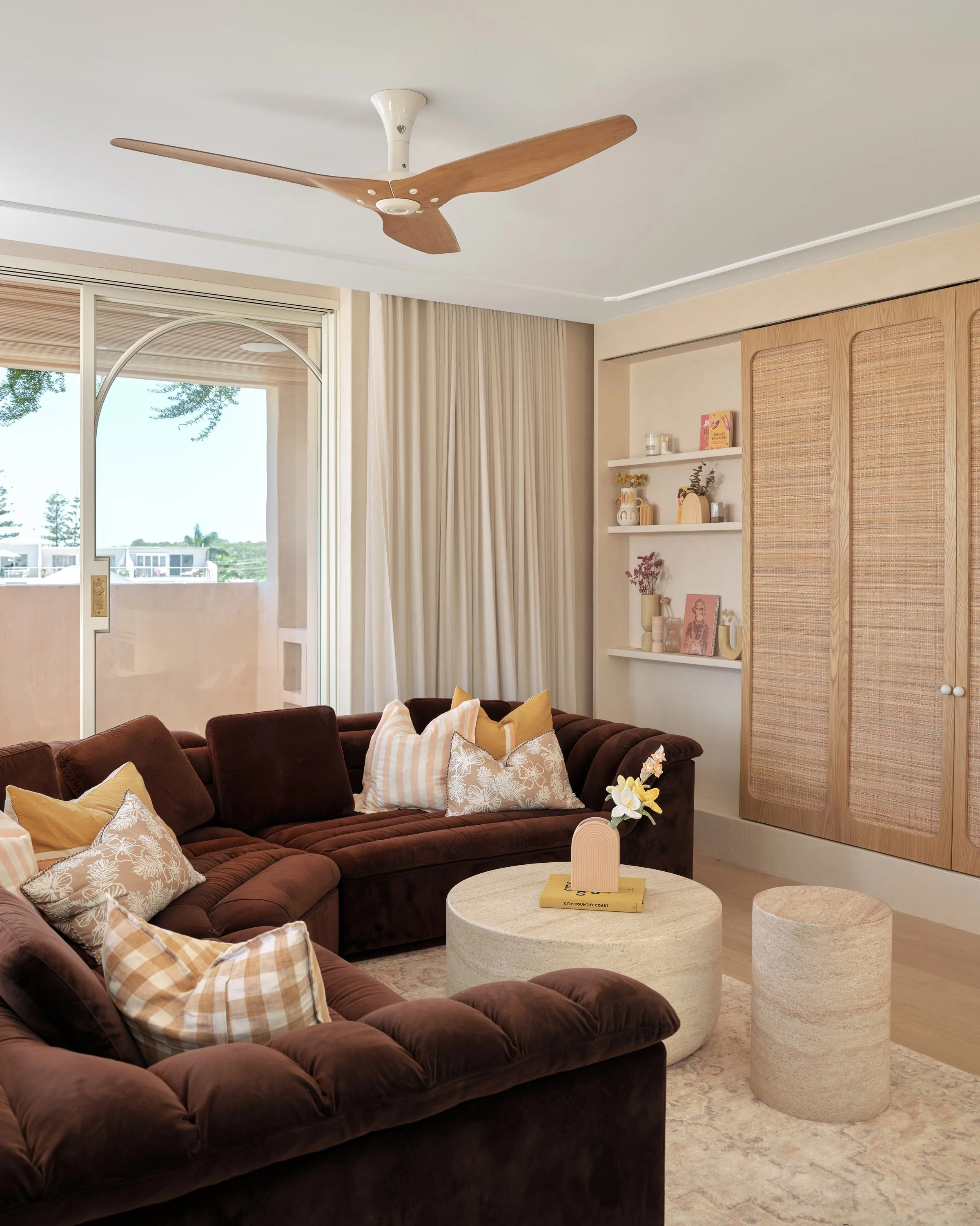 Living room with brown velvet sectional sofa, decorative pillows, beige curtains, a wooden ceiling fan, white shelves with decor, rattan closet doors, and a glass sliding door to a balcony.