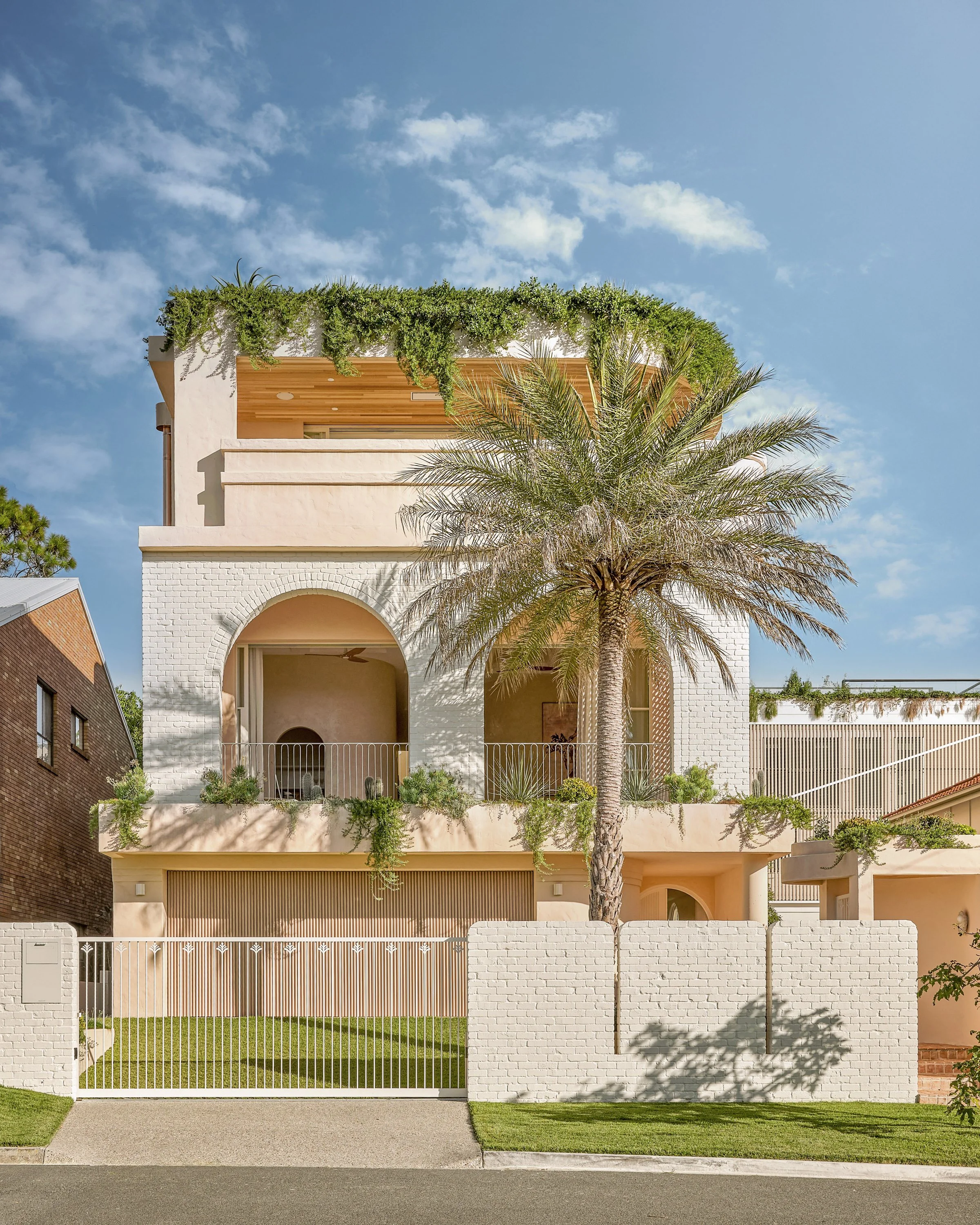 A modern multi-level house with pink and white exterior, large palm tree in front, green plants on balconies, white brick fence, and a clear blue sky.