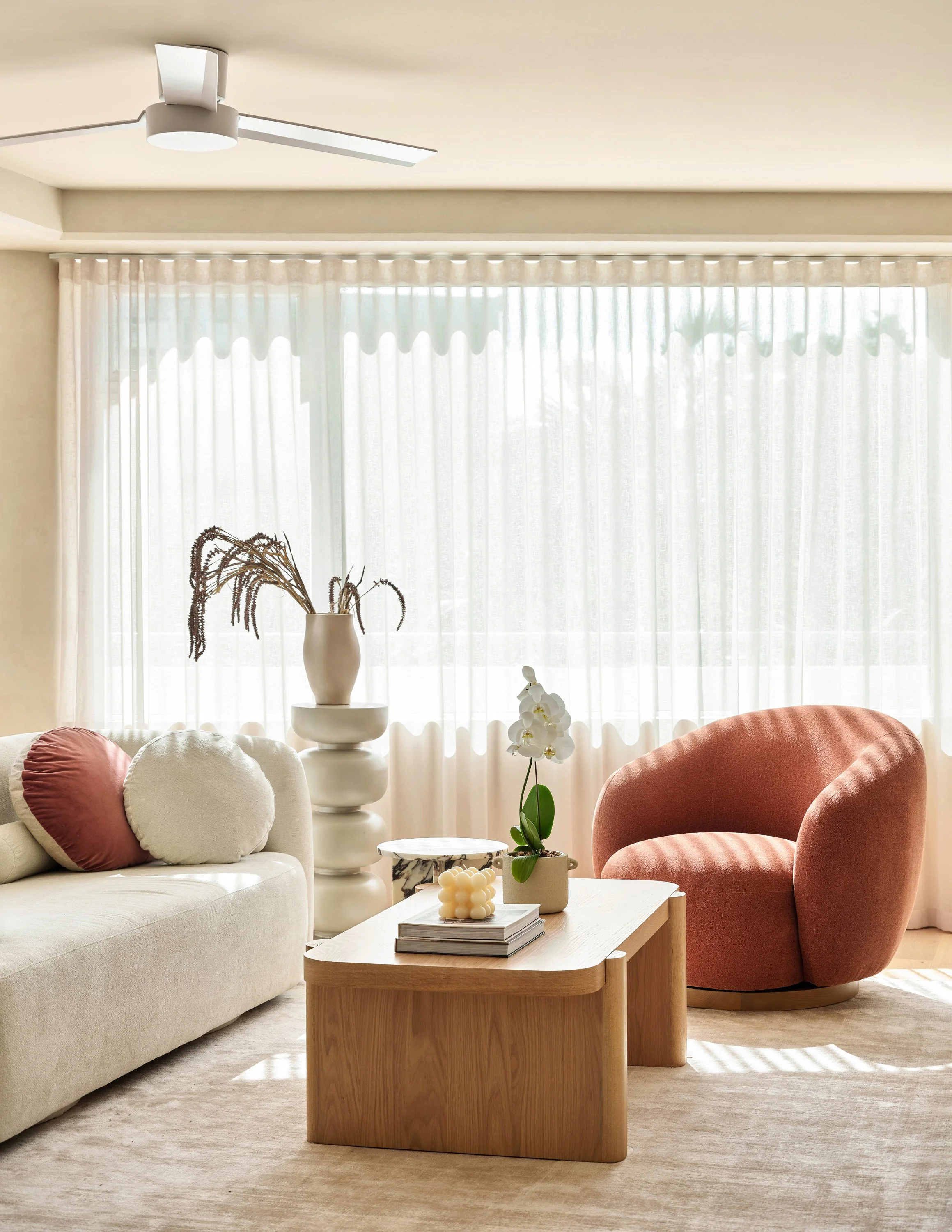 Living room with cream sofa, pink armchair, wooden coffee table, decorative pillows, potted orchid, modern vase with dried plants, and patterned lamp, with sheer curtains letting in natural light.