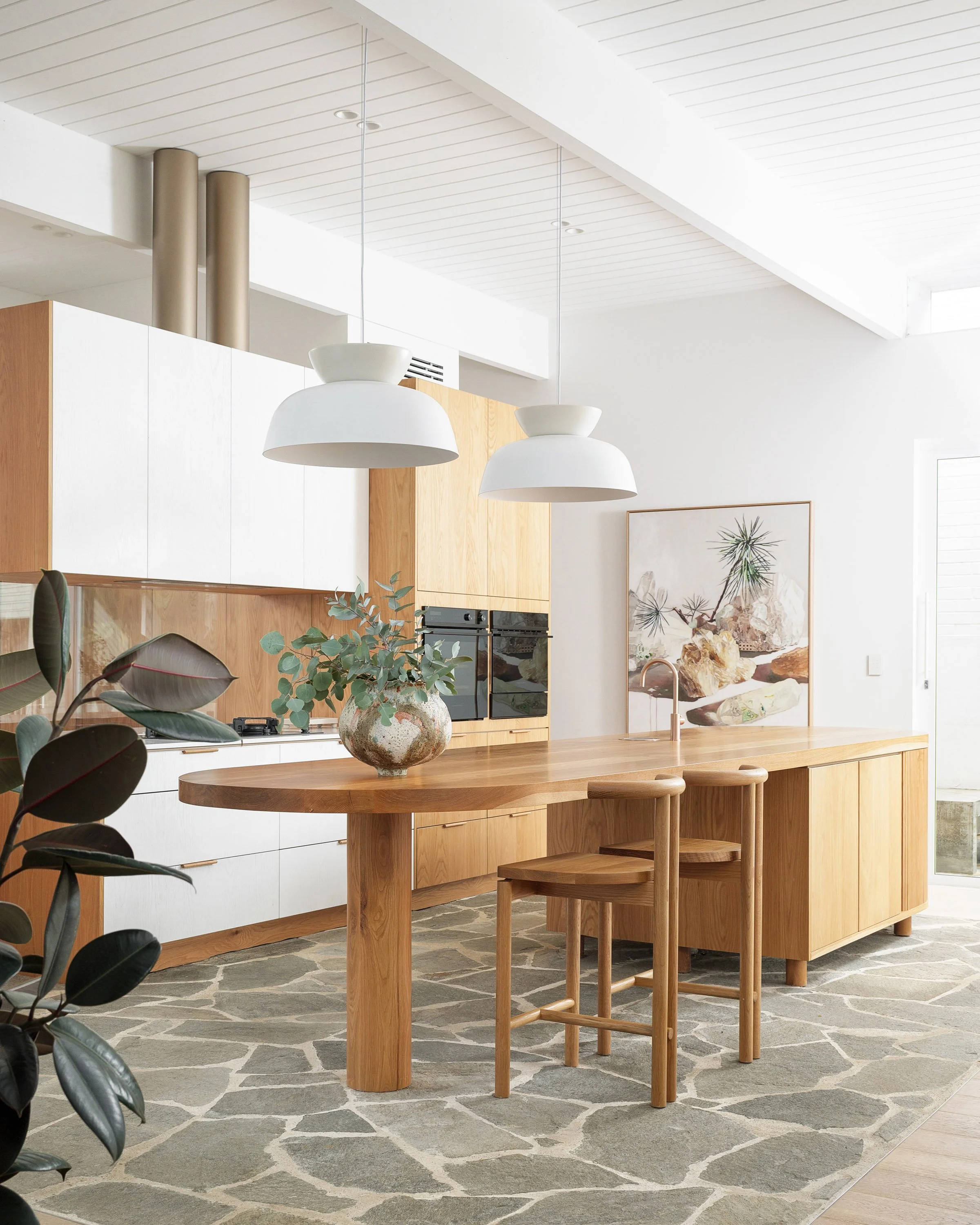 Modern kitchen with wooden cabinets and islands, two white hanging pendant lights, a ceramic vase with greenery on the island, a large framed artwork, and a patterned stone rug.