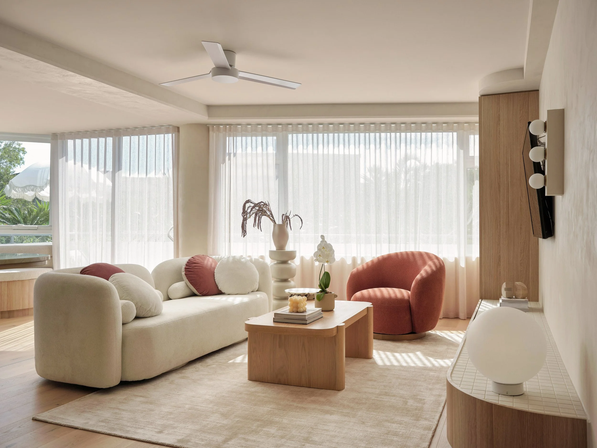 Living room with cream-colored sofa, pink and white cushions, wooden coffee table, pink armchair, and large window with sheer curtains