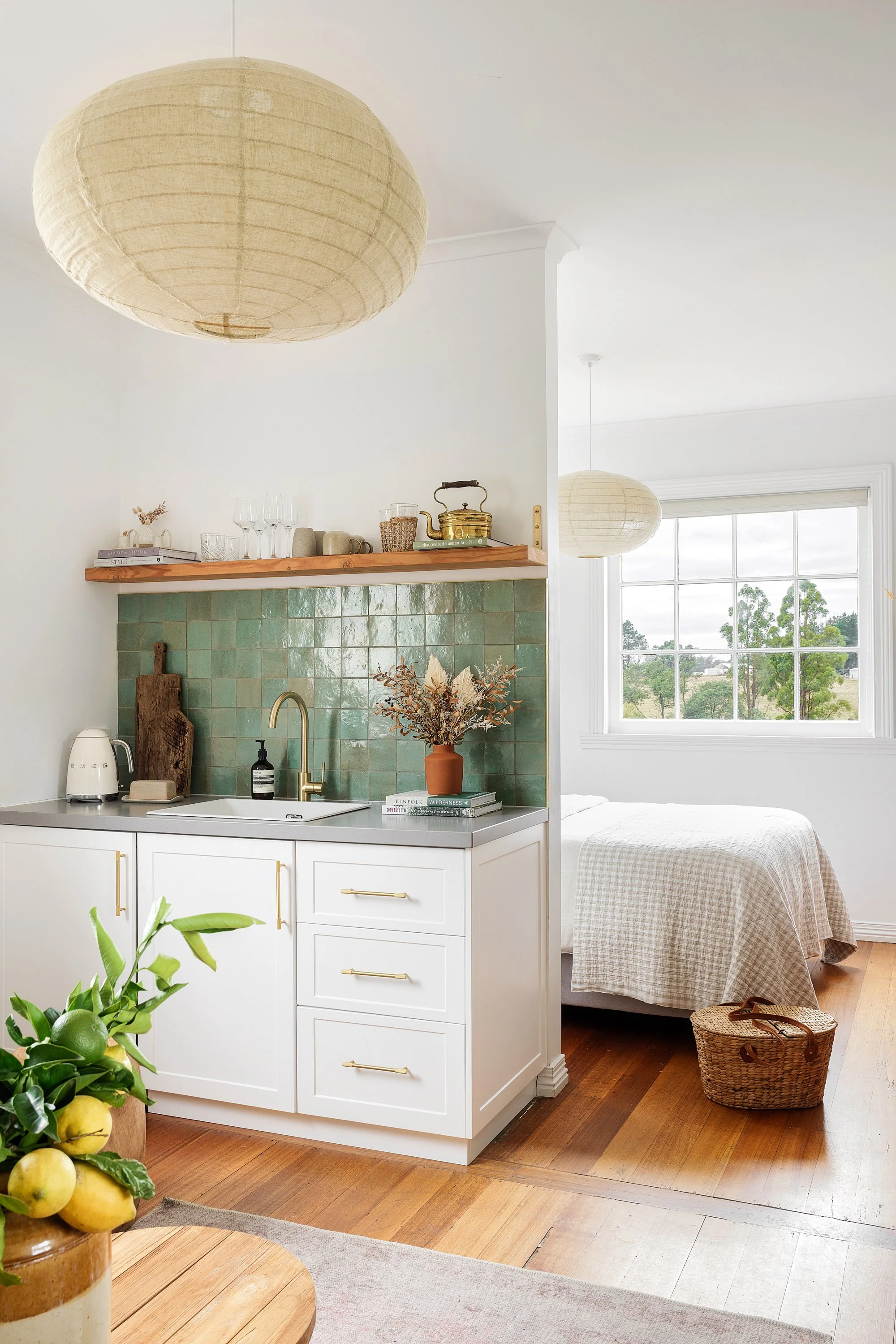Interior view of a small kitchen adjacent to a bedroom, featuring a white countertop, green tiled backsplash, and various decorative items including a vase with dried flowers, books, and kitchenware, with a window showing trees outside.