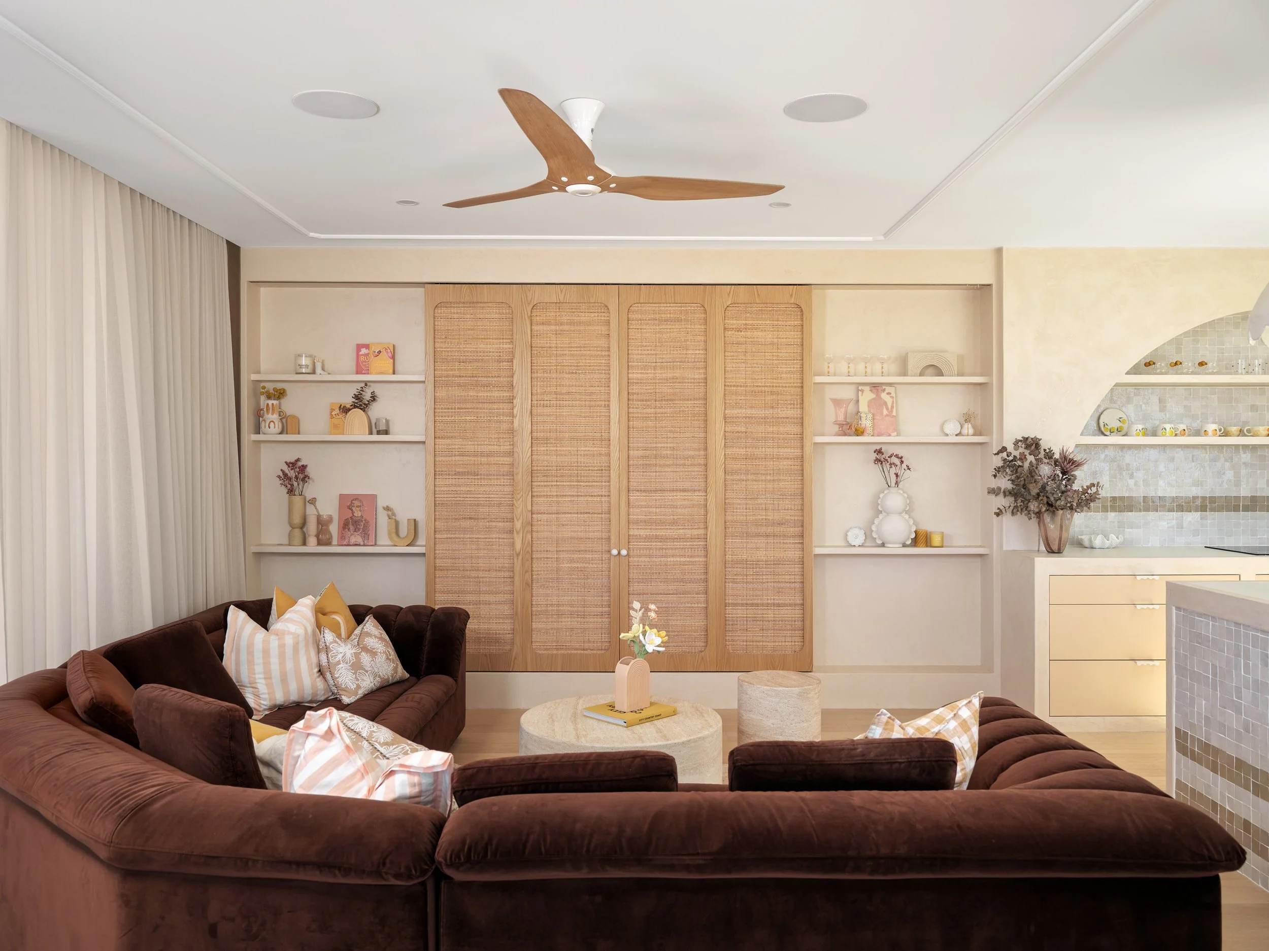 Living room with brown velvet sectional sofa, pillows, round coffee table, beige built-in shelves with decorative objects, ceiling fan, and partial view of kitchen area.