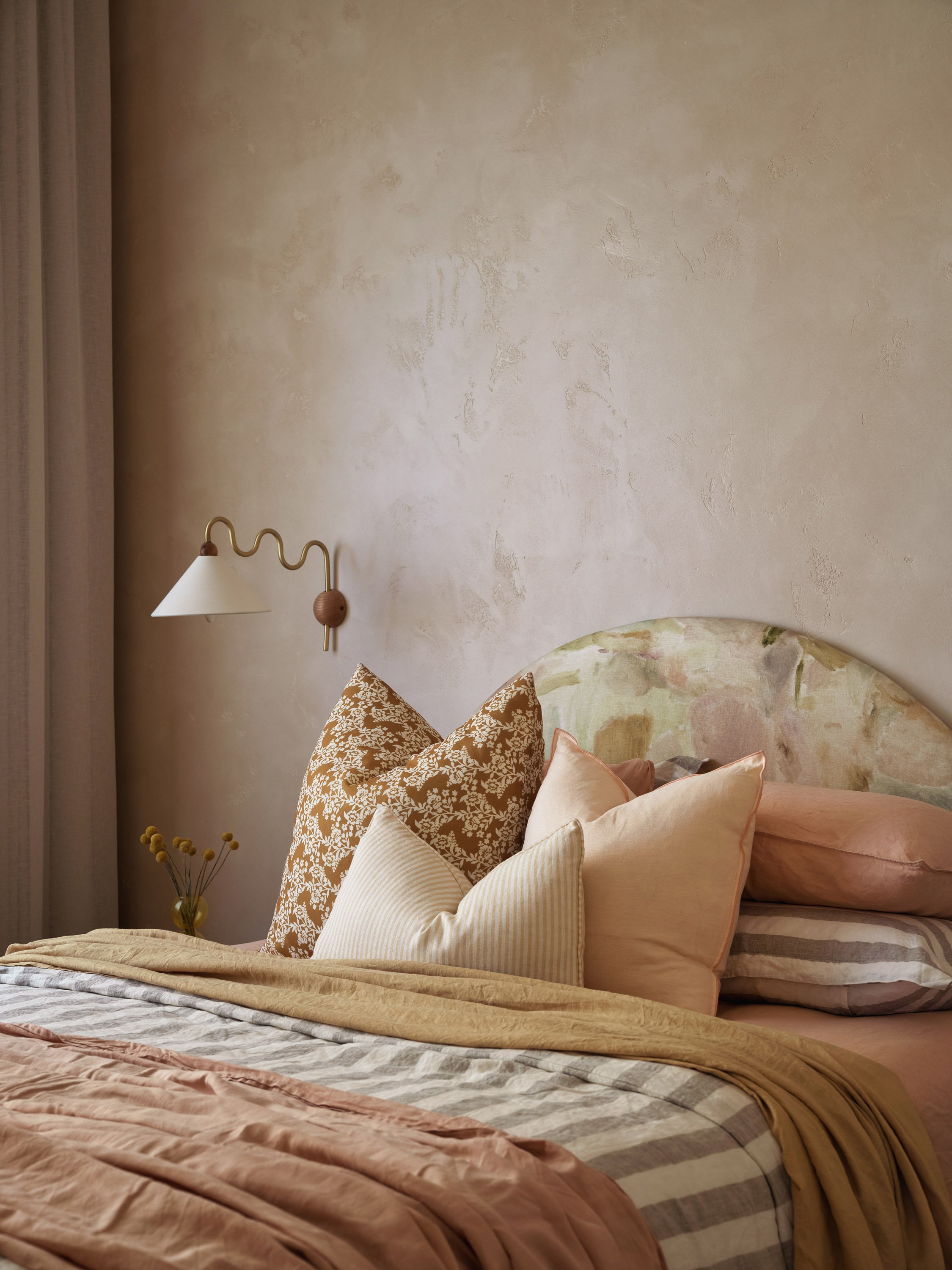 A cozy bedroom with a bed featuring multiple beige, floral, and striped pillows, a mix of beige, pink, and gray bedding, a textured beige wall, a sconce light on the wall, and a small vase with yellow flowers on a nightstand.