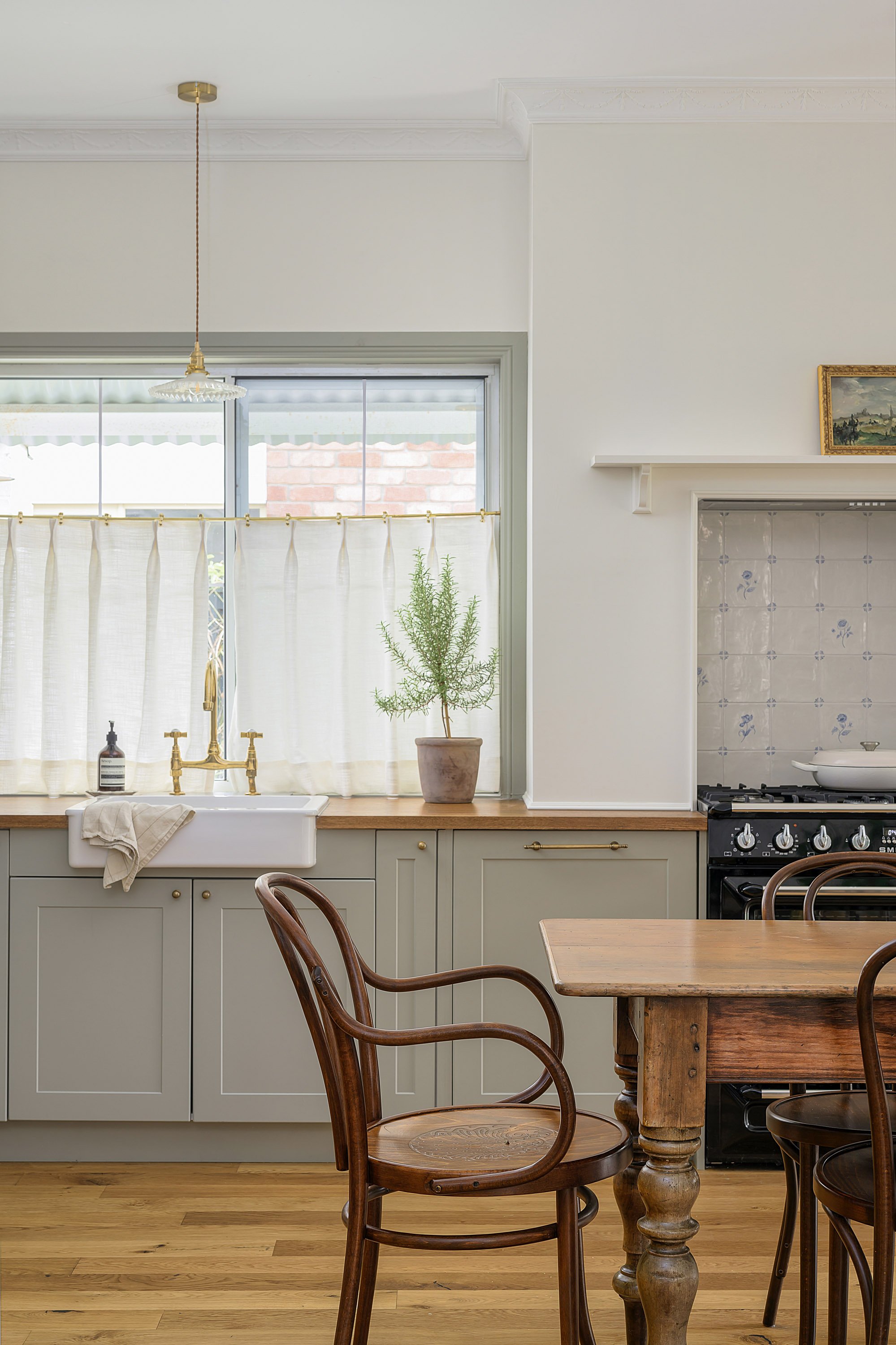 A cozy kitchen featuring light green cabinetry, a white ceramic farmhouse sink, a window with white curtains, a potted plant on the window sill, a wooden dining table, and wooden chairs.
