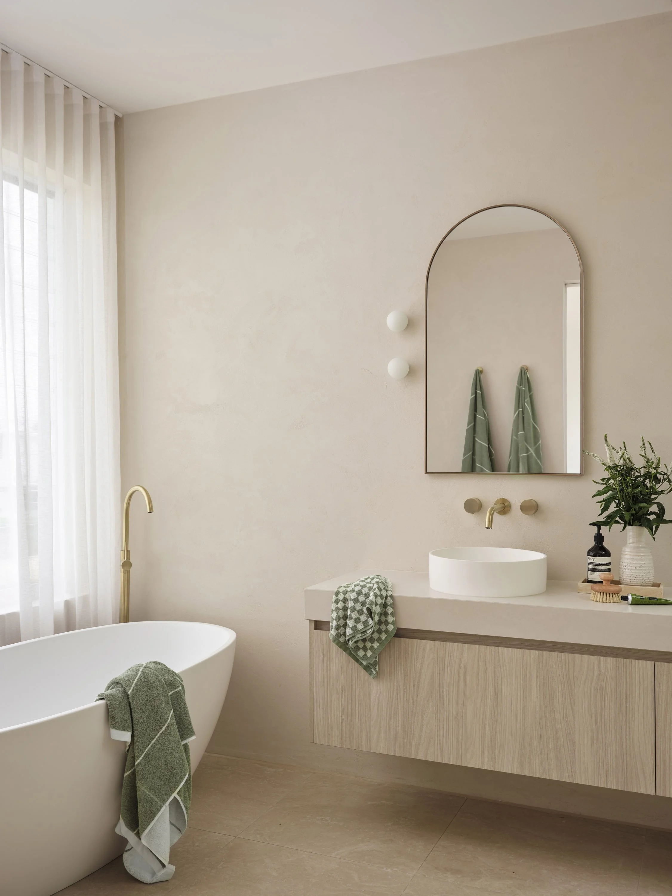 Modern bathroom with a free-standing bathtub, a floating vanity with a vessel sink, a large arched mirror, green towels, a potted plant, and neutral-colored decor with natural light from a window with sheer curtains.