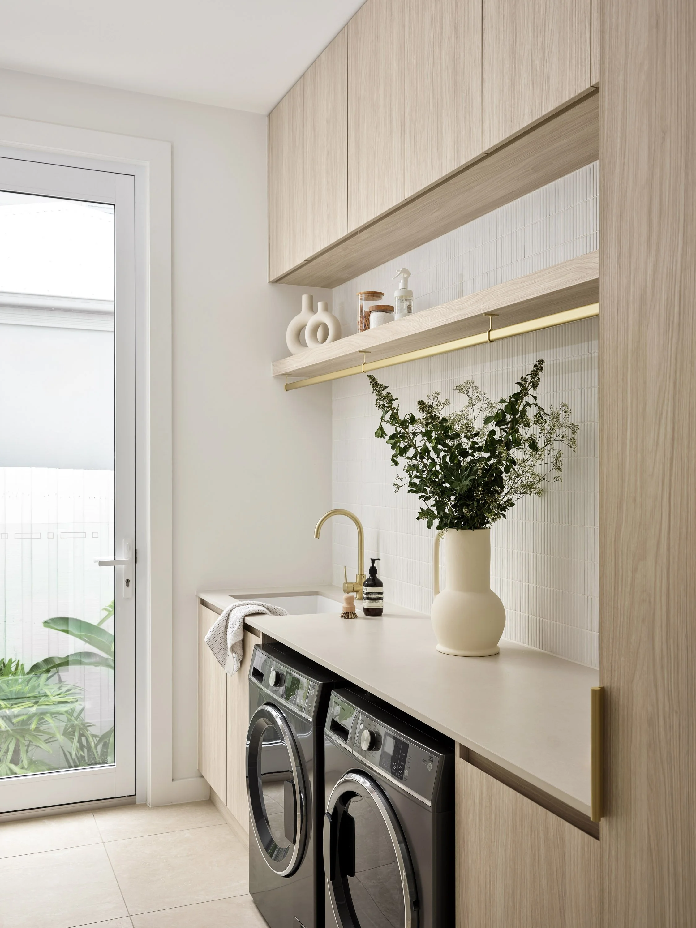 Modern laundry room with a white countertop, a vase with greenery, and front-loading washer and dryer, with wooden cabinets and shelf above, near a glass door with plants outside.