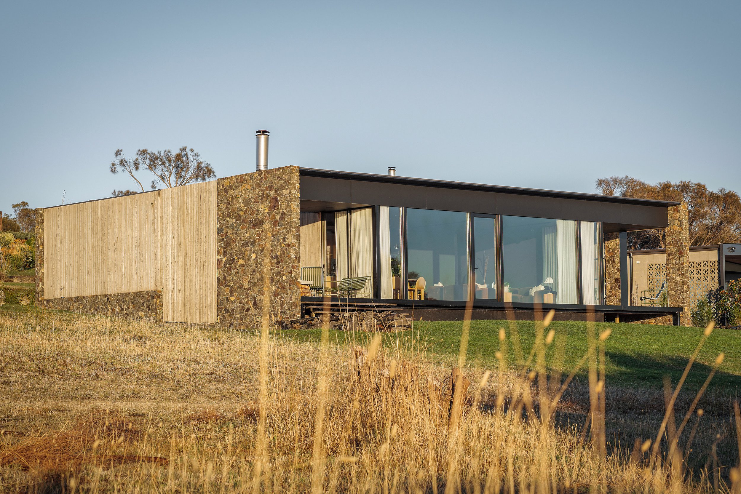 Modern house with stone and wood exterior, large glass windows, sitting on a grassy hill with dry grass in the foreground.