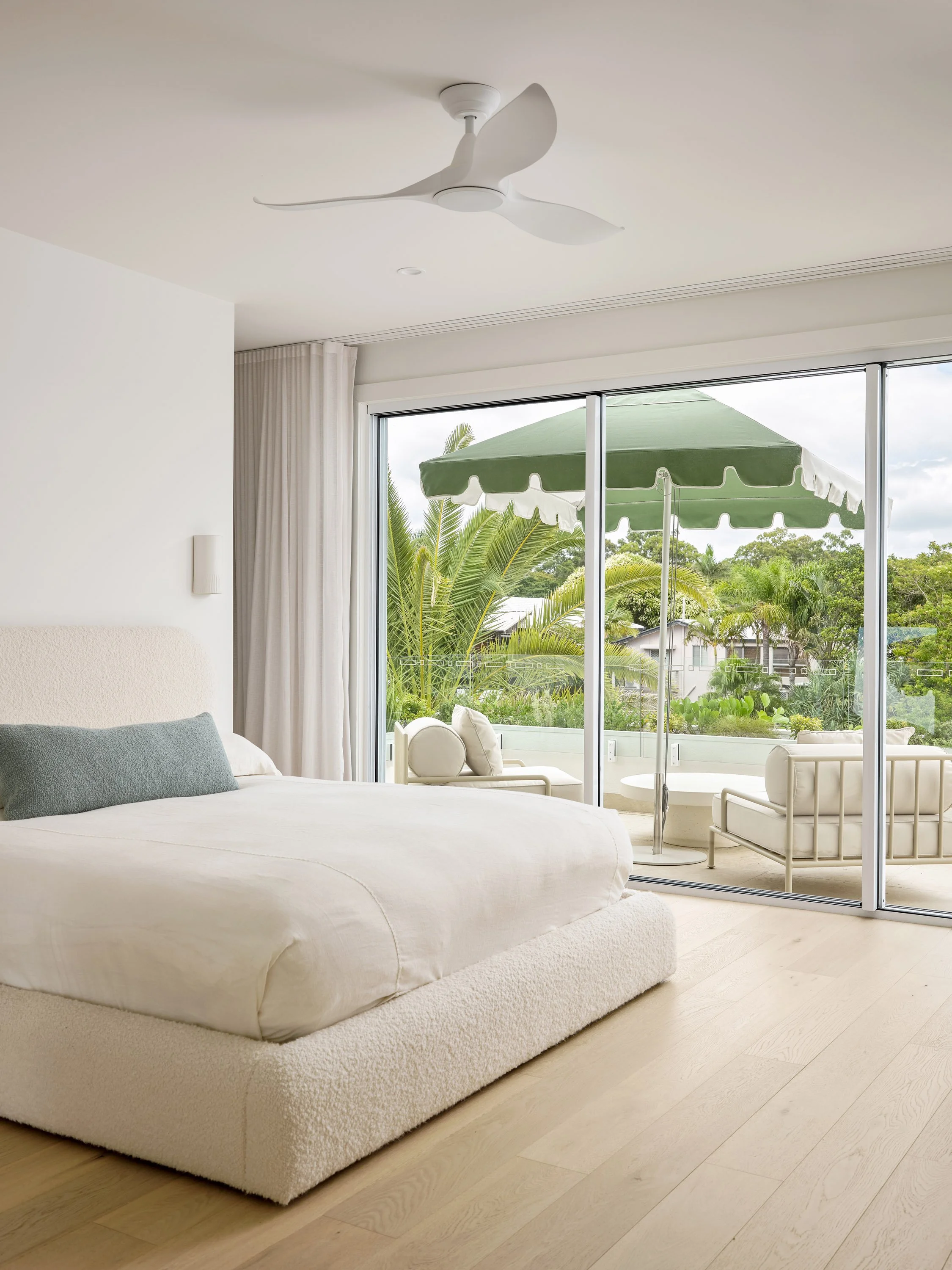 Bedroom with a large bed, sliding glass doors leading to a balcony with outdoor seating, green umbrella, tropical plants, and trees outside.