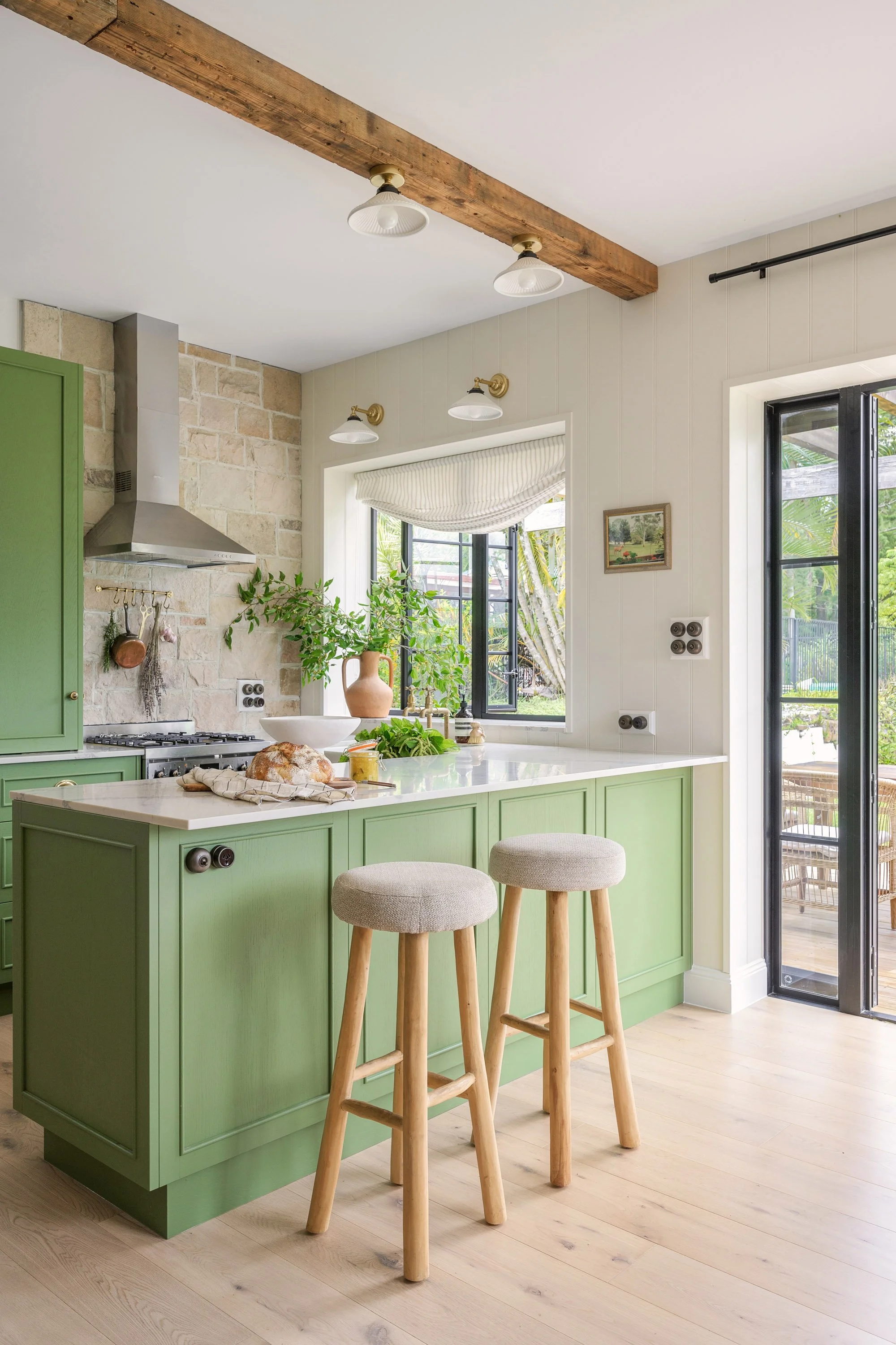 Bright kitchen with green cabinetry, a white countertop island with two stools, a window with a view of a garden, and a door leading outside.
