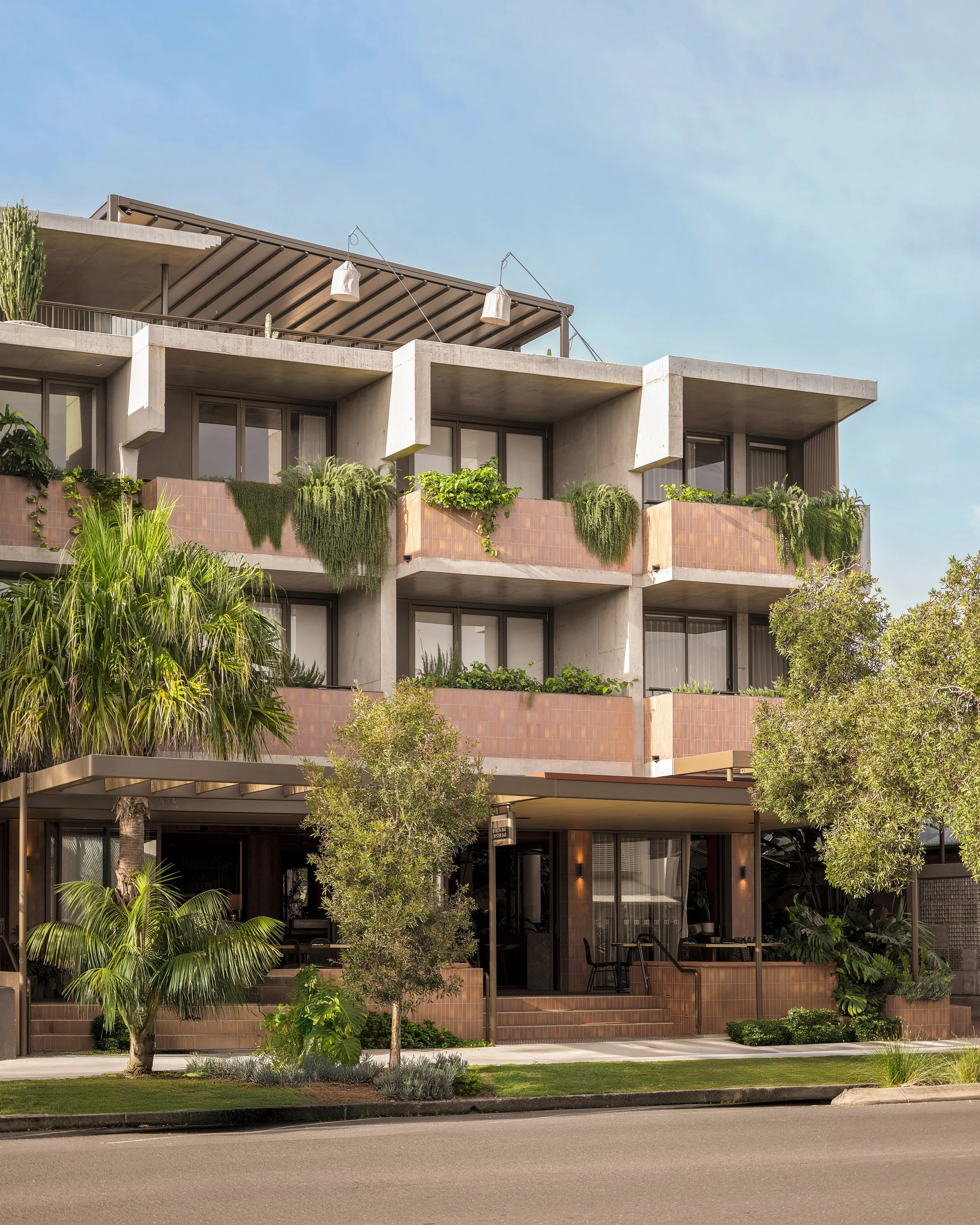 Modern multi-story residential building with balconies, lush greenery, and trees in the foreground under a clear blue sky.