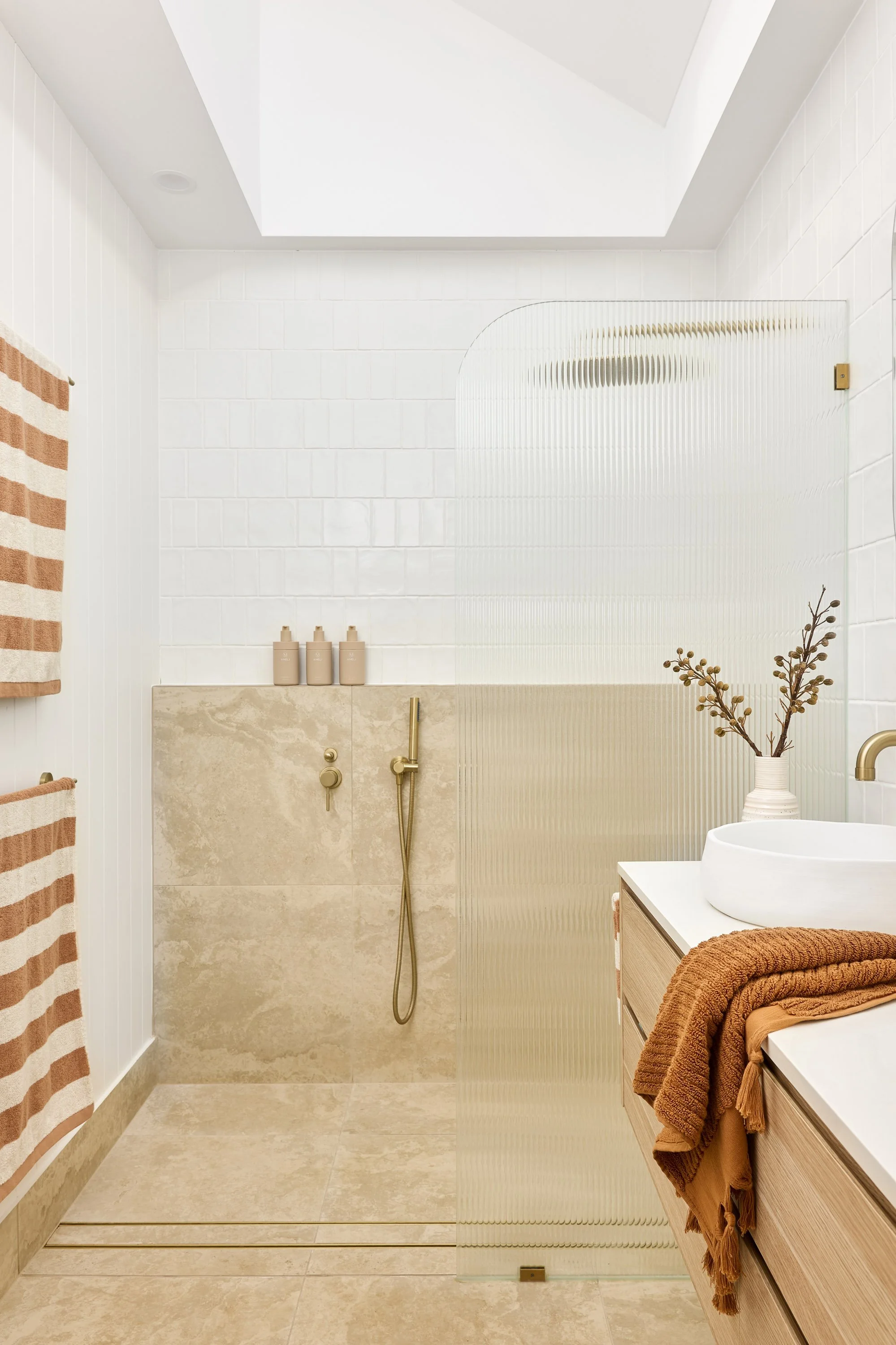 Modern bathroom with beige tiled floor, white tiled walls, a skylight ceiling, and a glass shower divider. There are beige and white striped towels on a wall rack, a vanity with a white sink, a decorative vase with dried branches, and beige toiletrie