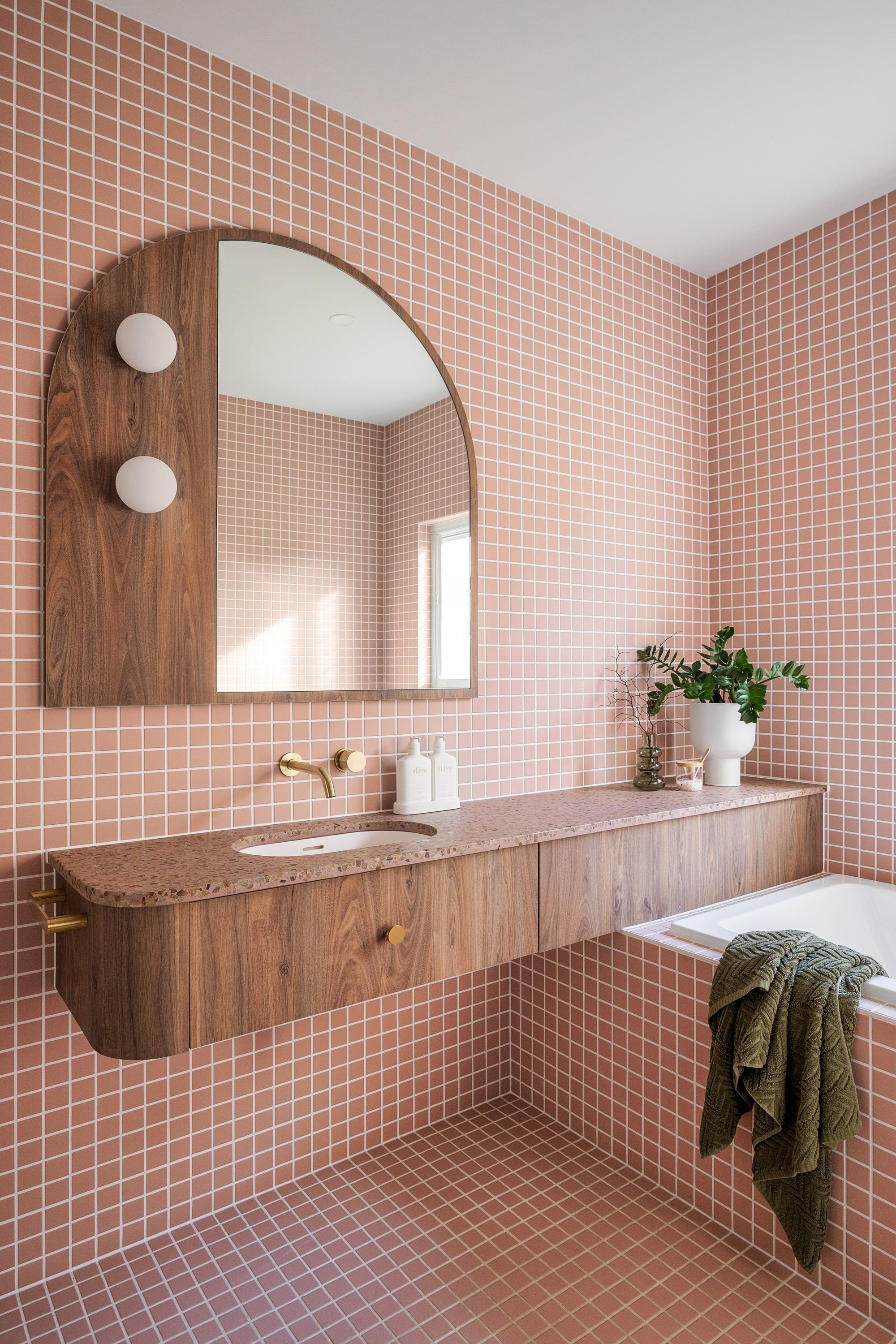 A modern bathroom with pink mosaic tile walls and floor, a wooden vanity with a pink countertop, a clip mirror above it, white bottles, and a bathtub with a green towel hanging over its edge, decorated with a potted plant and vases.