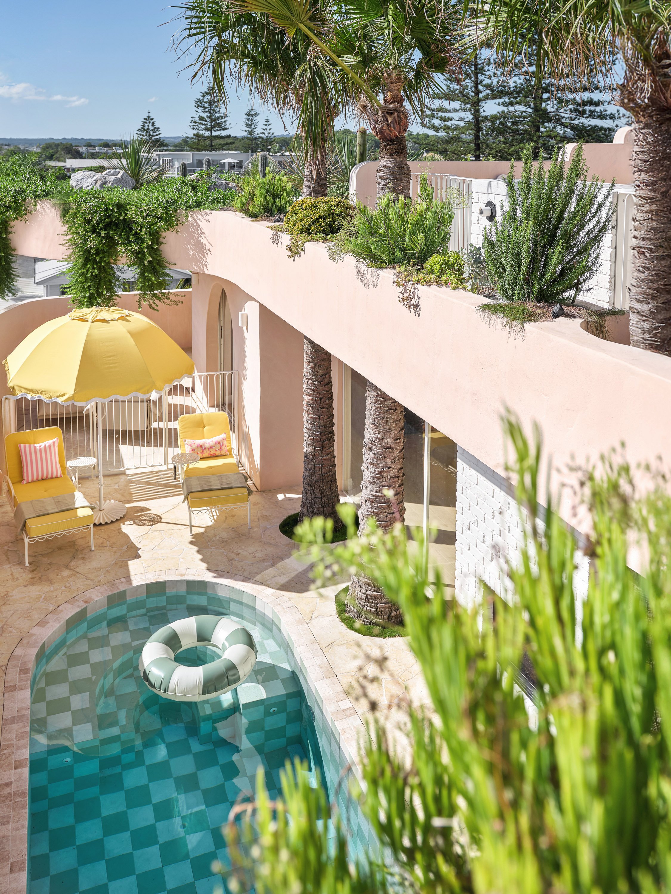 A sunny outdoor patio with a swimming pool, yellow chairs with pink cushions, a yellow parasol, lush greenery, and tall palm trees.