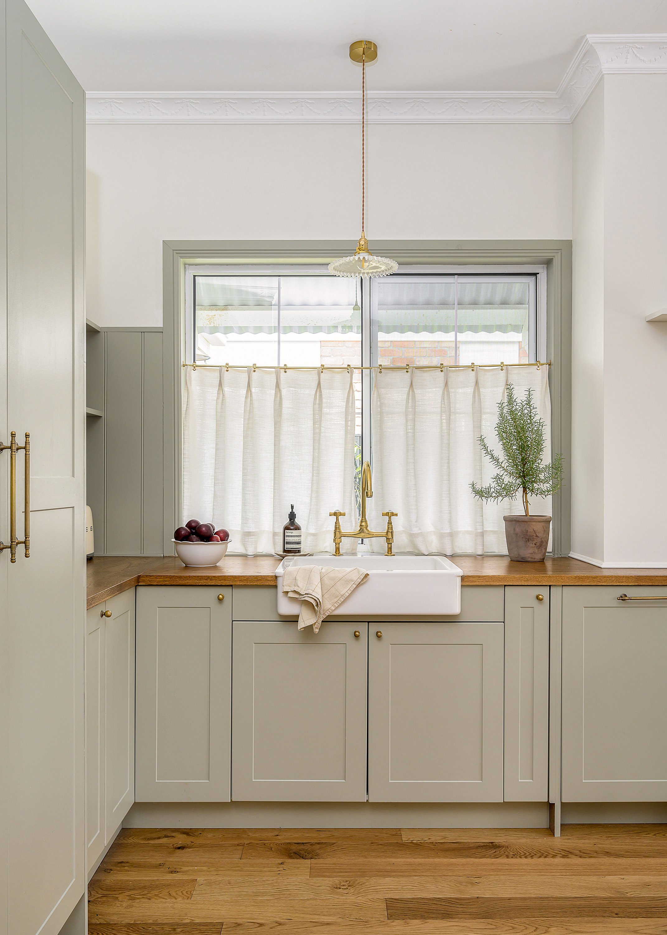 Kitchen with a farmhouse sink, beige cabinets with gold knobs, a window with white curtains, a bowl of purple apples, a soap dispenser, a potted plant, and hardwood floors.