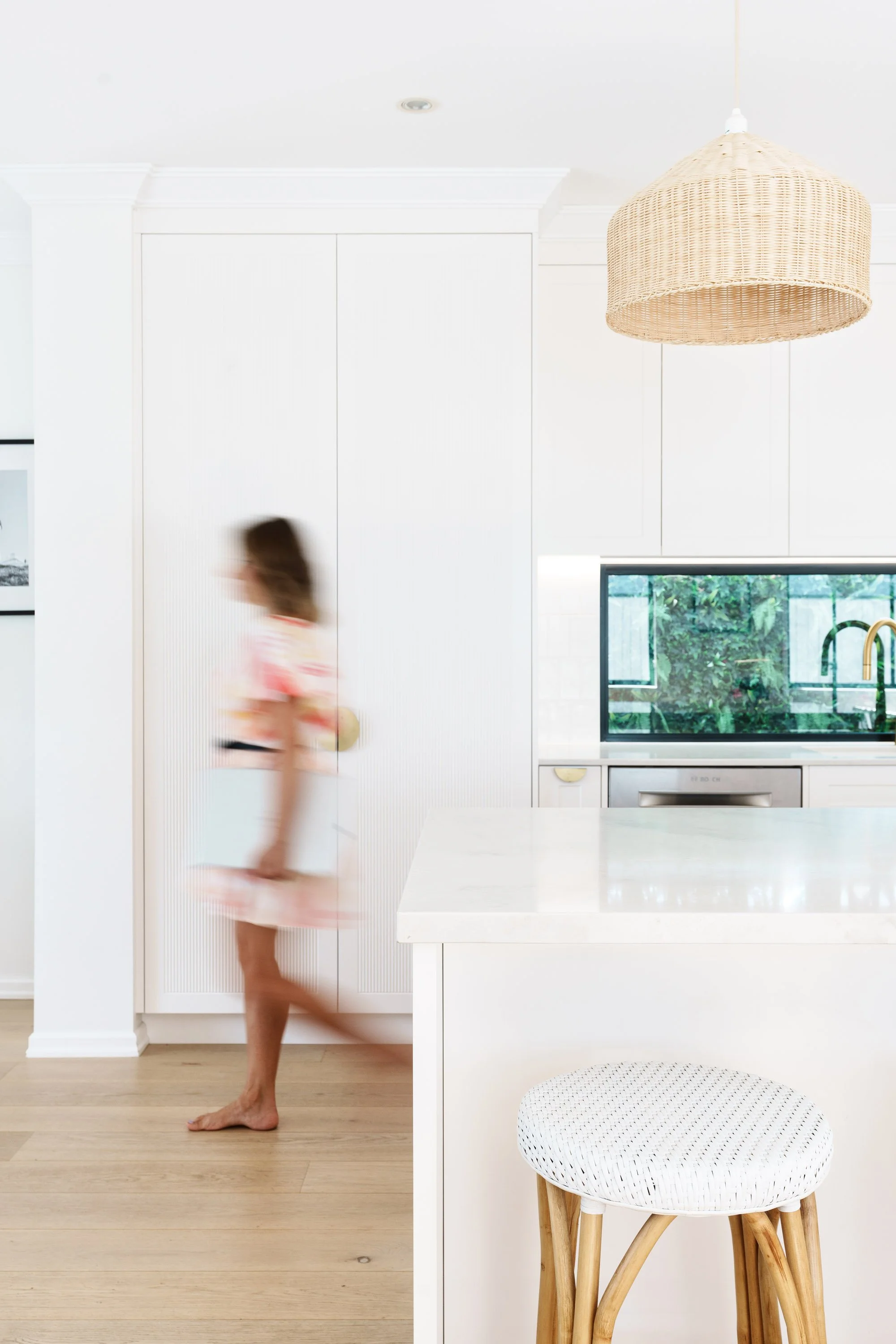 A blurred woman in a colorful dress walking in a modern, white kitchen with a white marble island, wooden stool, and greenery visible through the window.