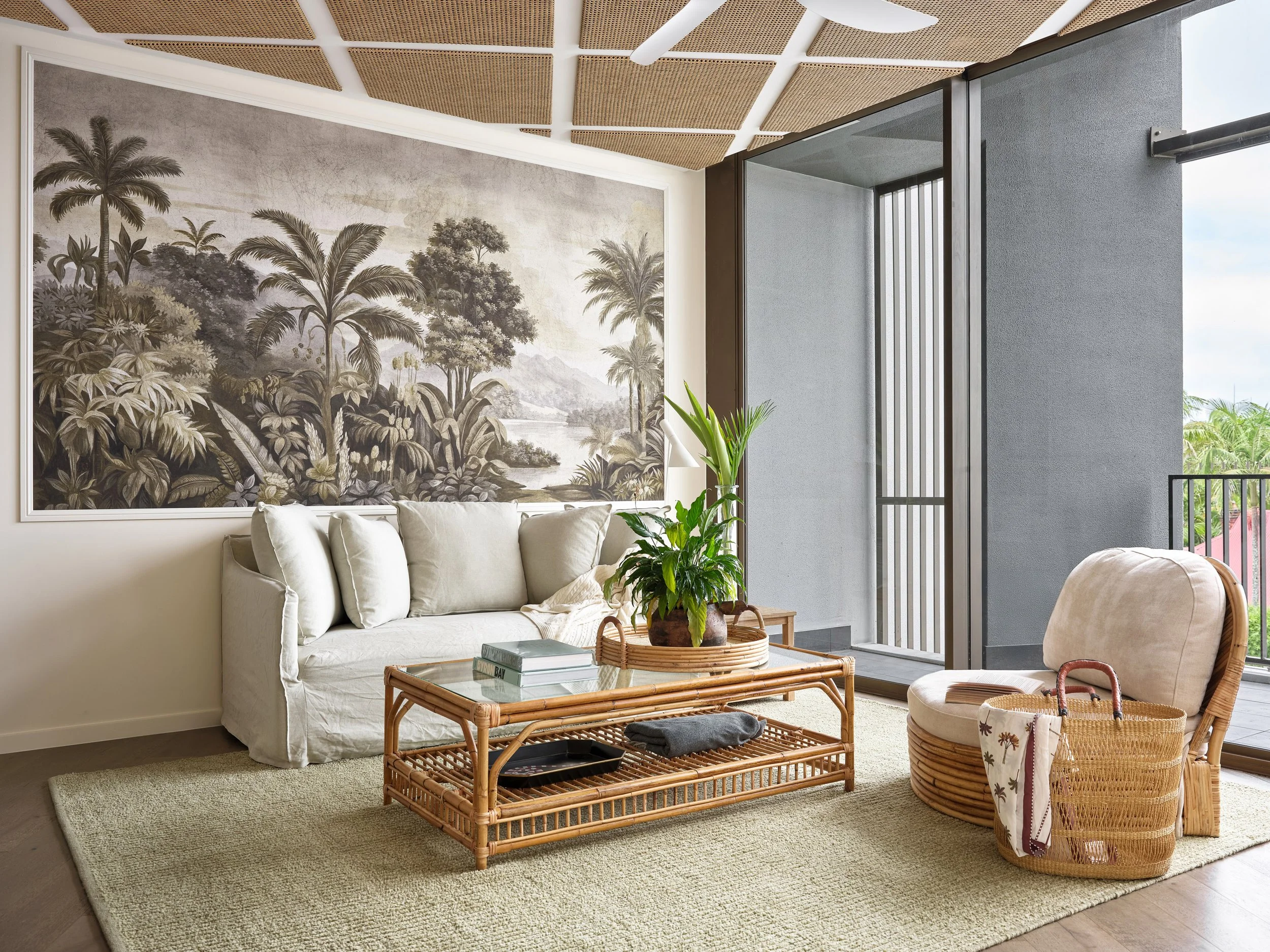 A cozy living room with a beige sofa, a wicker coffee table, and a round wicker chair. A large potted plant is on the table. The wall features a large black-and-white tropical landscape mural. An open balcony door shows greenery outside, and the ceil