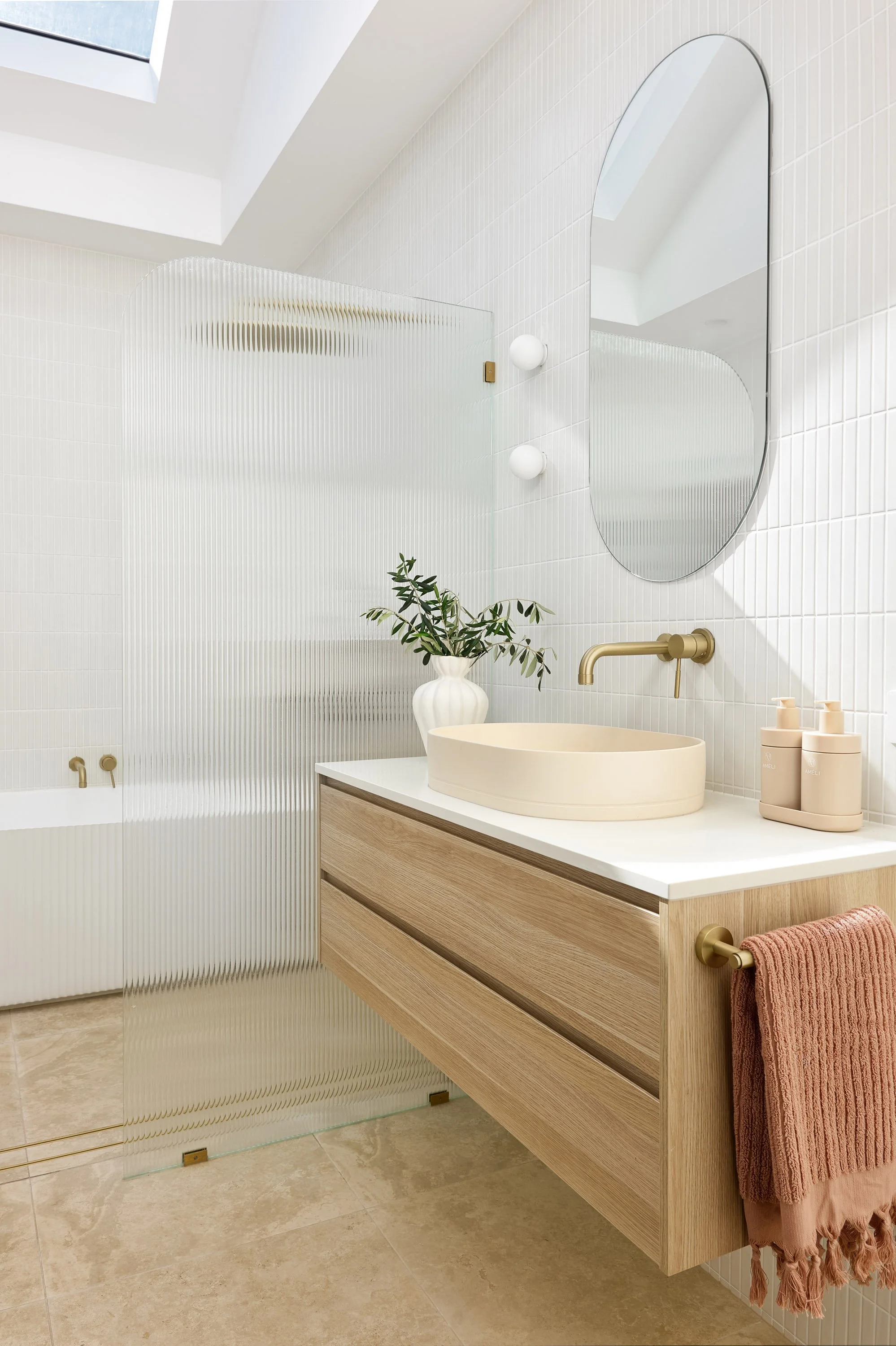 Modern bathroom with a floating wooden vanity, a beige countertop basin, a large oval mirror, a gold wall-mounted faucet, a potted plant, and lotion bottles, with a skylight ceiling.