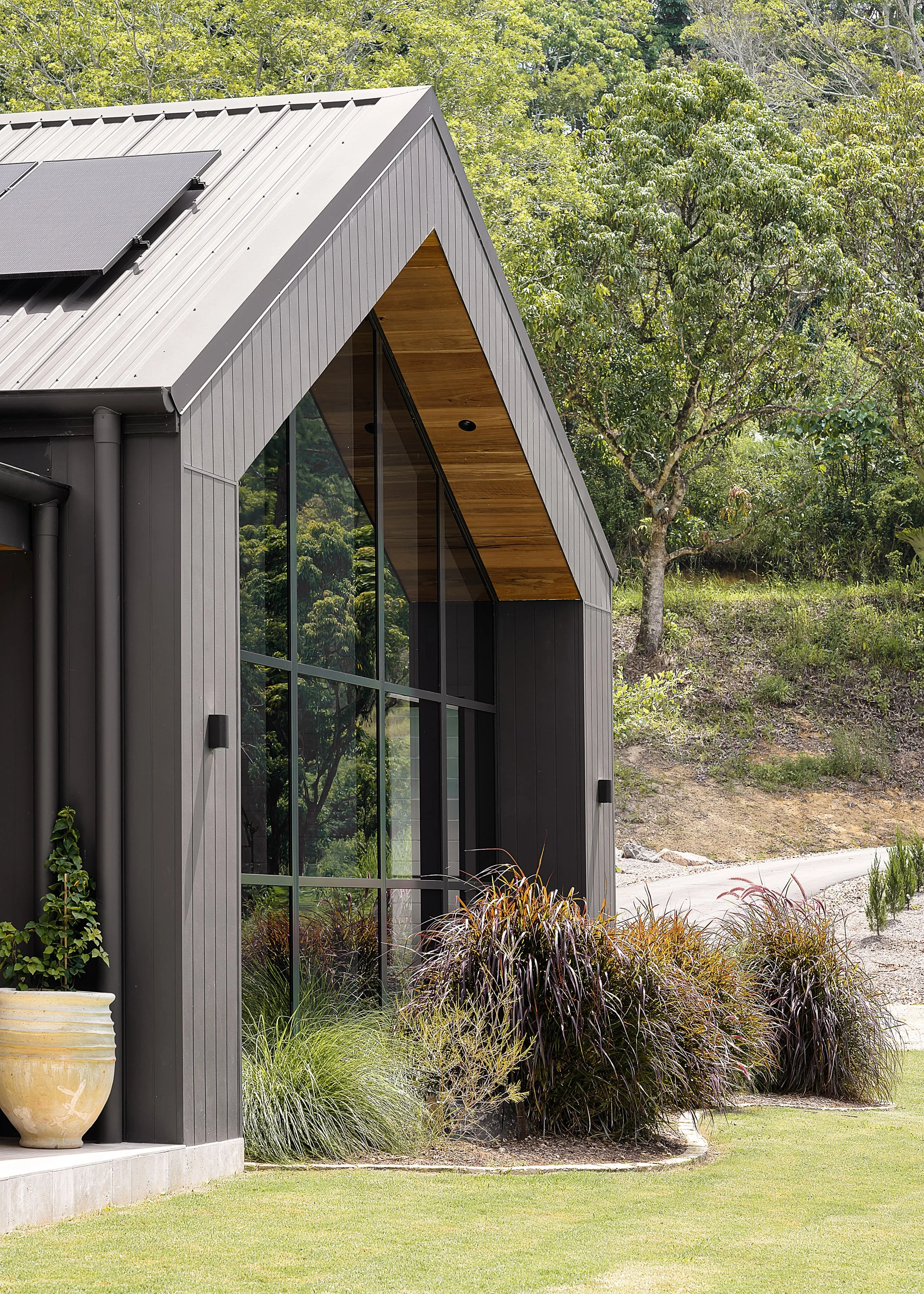 Modern house with black exterior, large glass windows, sloped metal roof, and surrounding landscaped garden with ornamental grasses and potted plants.