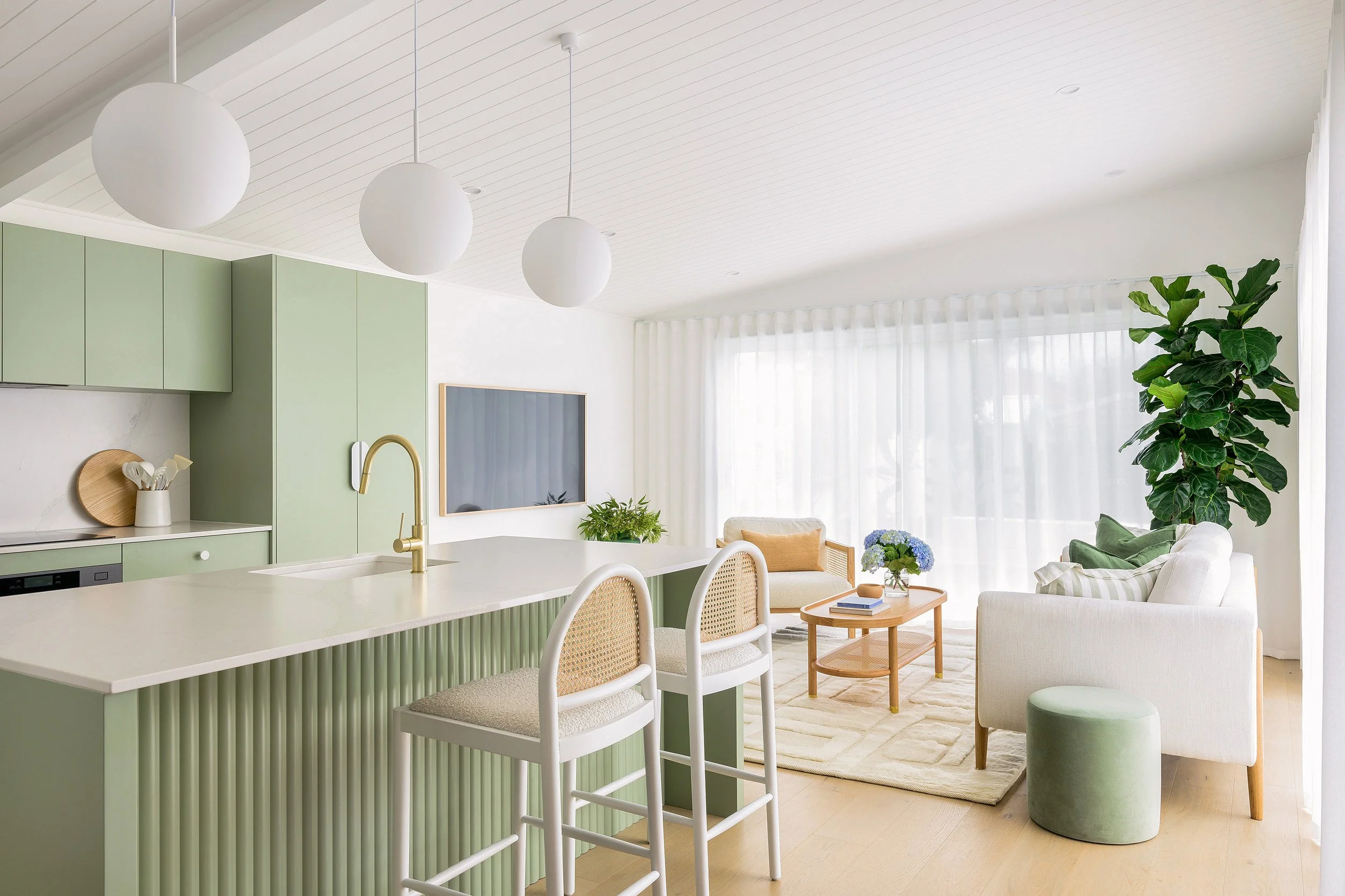 Bright living room with white and pastel green kitchen island, bar stools, a white sofa, coffee table, tall leafy plant, and sheer curtains