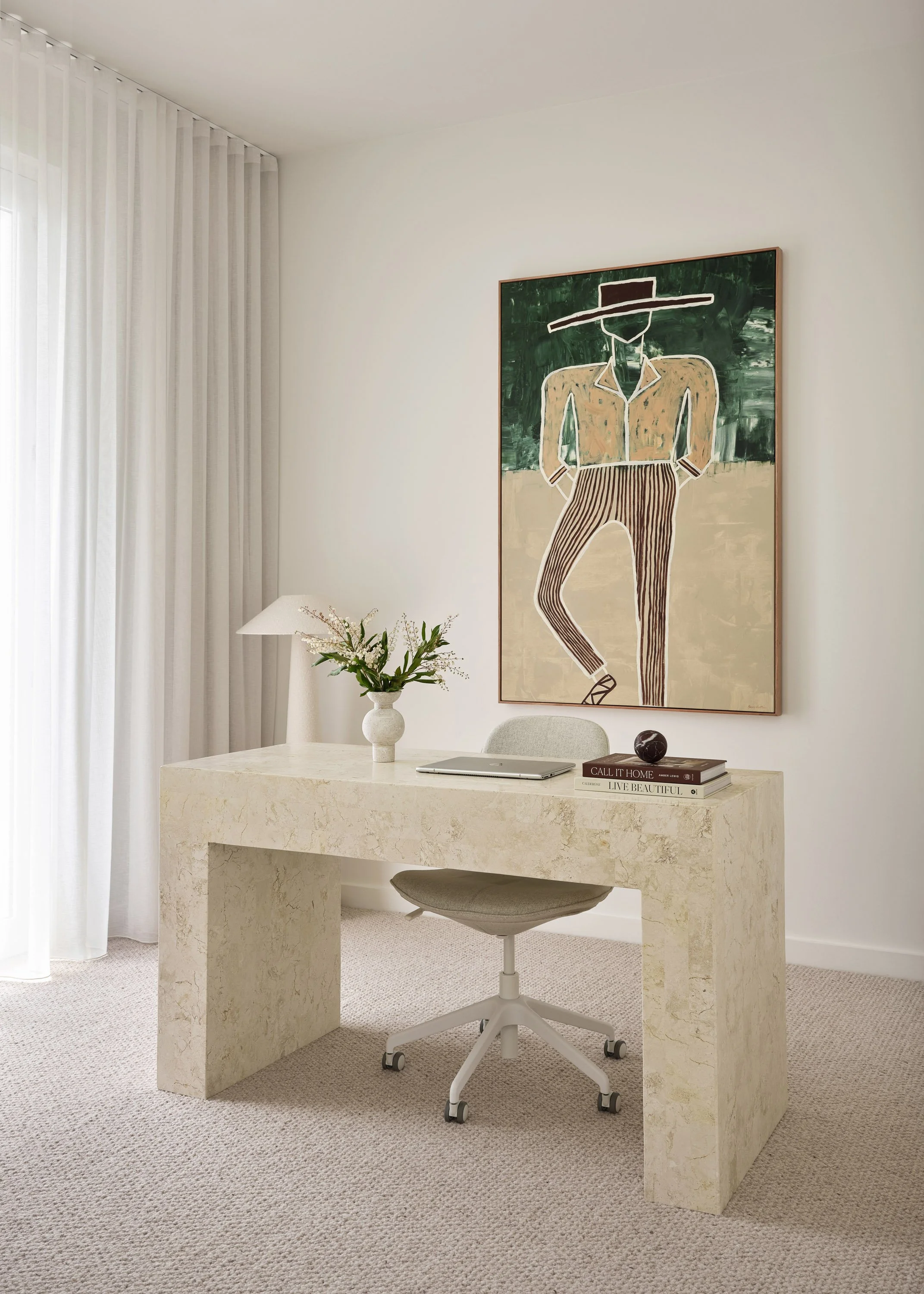 A simple modern office space with a marble desk, a gray office chair, a white vase with flowers, a table lamp, some books, and a large abstract painting of a person with a wide-brimmed hat hanging on the wall behind the desk. The room has white walls