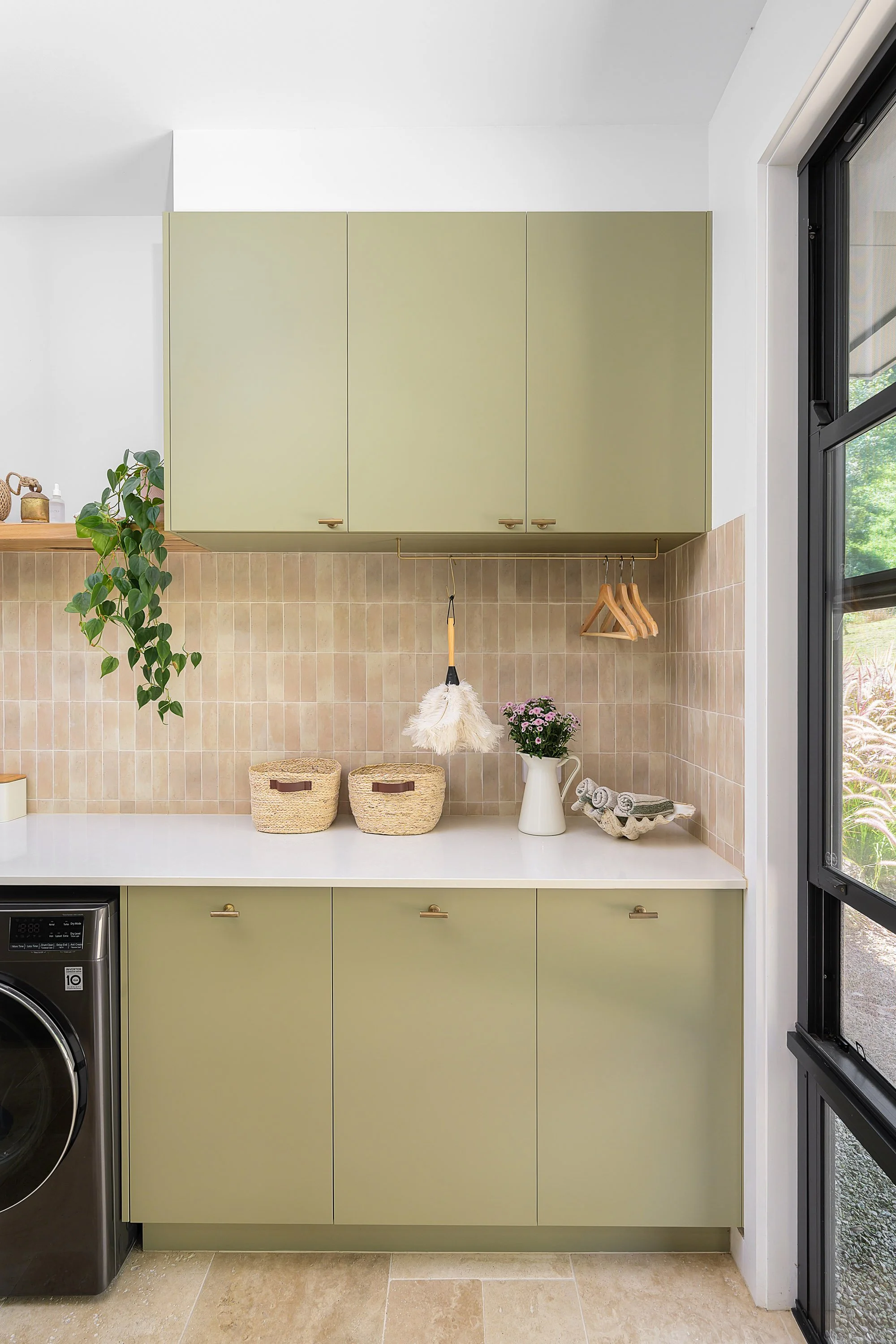 A modern laundry room with green cabinets, beige tiled backsplash, white countertop, and a window on the right side showing greenery outside. Decor items include baskets, a potted plant, towels, and a hanging duster.