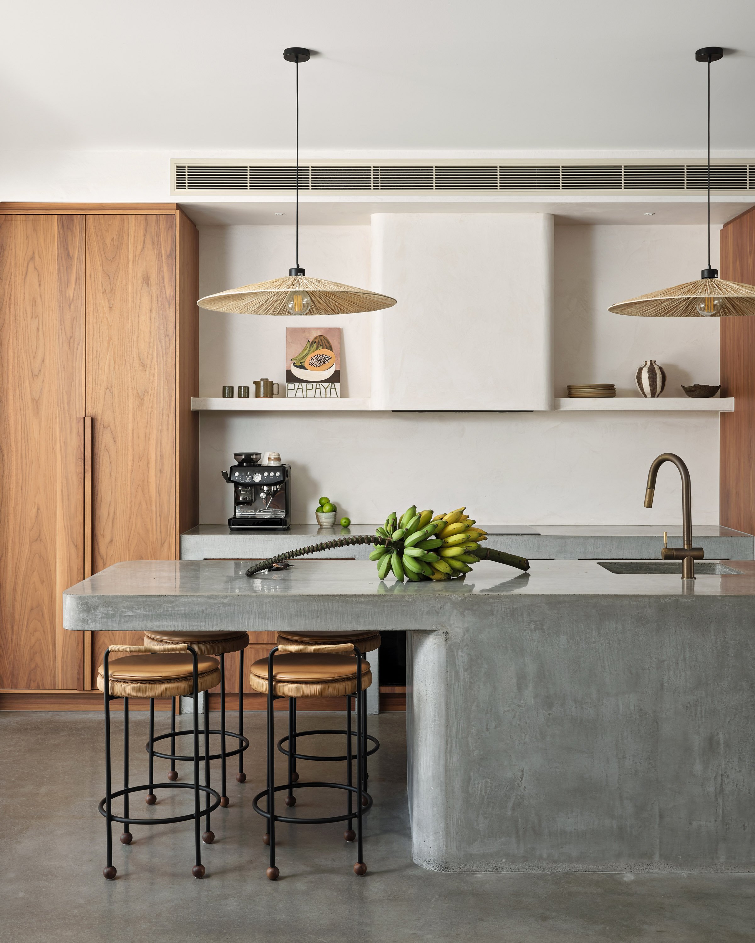 Modern kitchen with concrete island, hanging pendant lights, wooden cabinets, banana bunch on the counter, and a coffee maker.