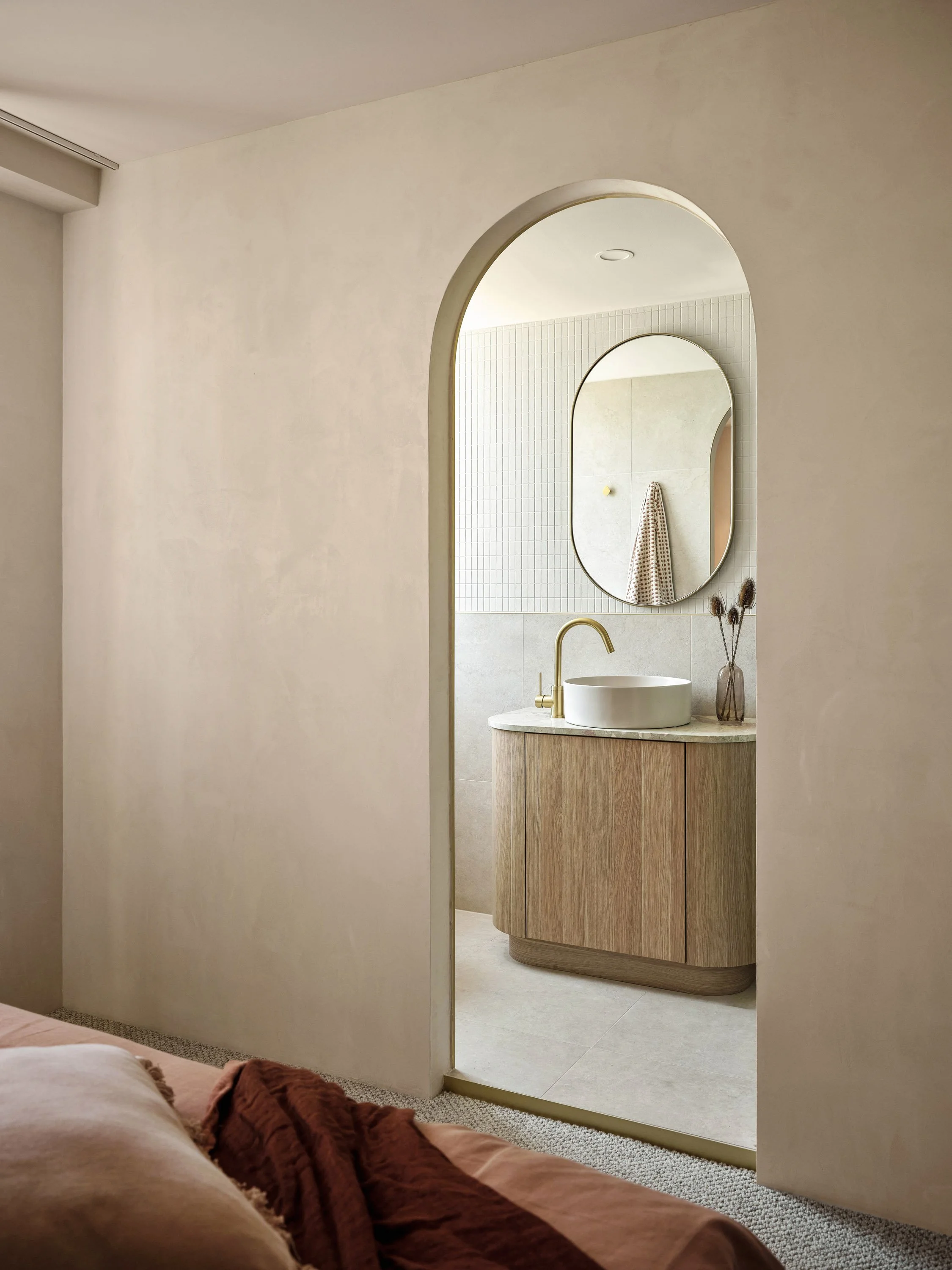 A view of a modern bathroom seen through an arched doorway, featuring a round vessel sink with a gold faucet, a mirror on the wall, a vase with dried flowers, and a wooden cabinet.