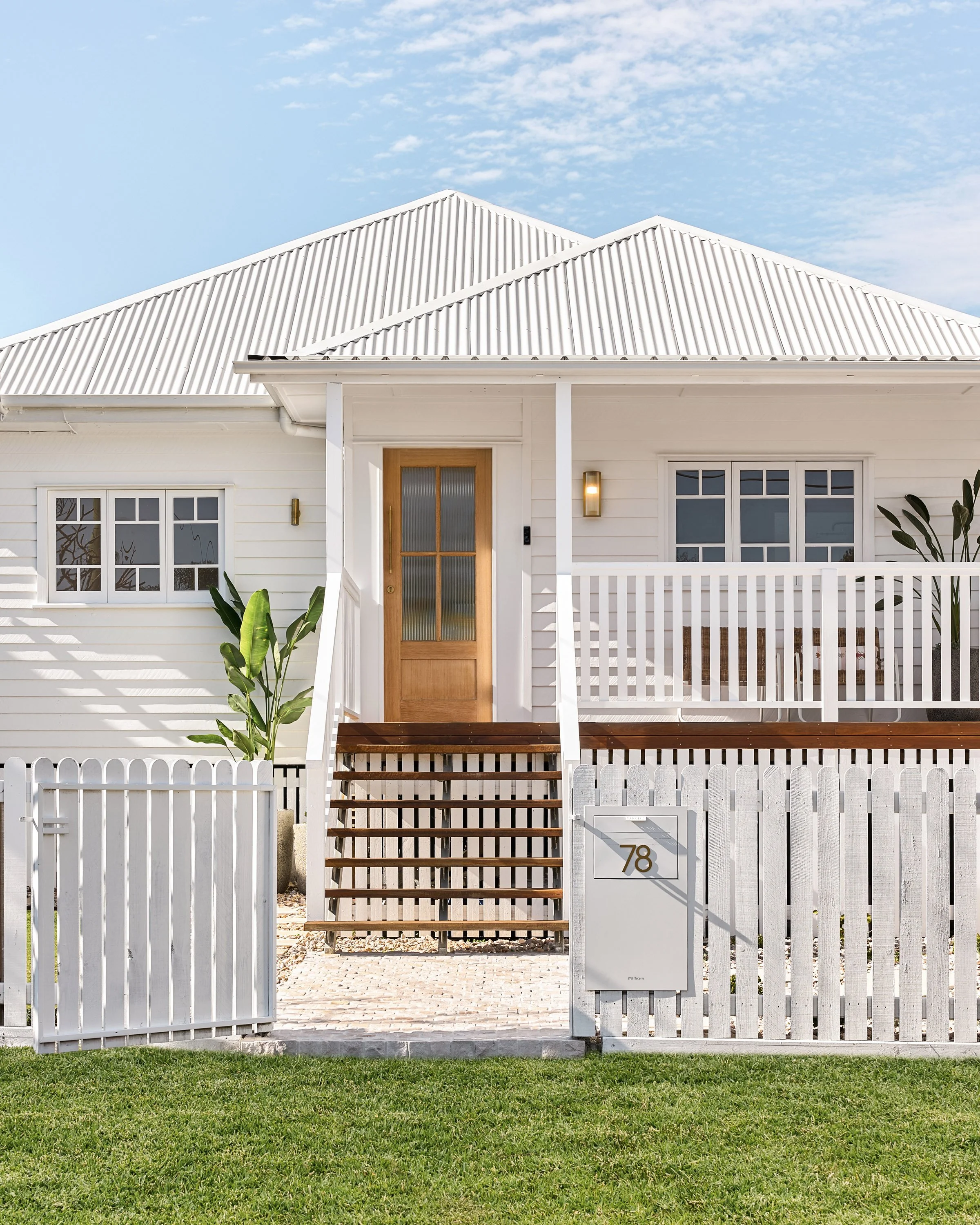 White house with a metal roof, wooden front door, front porch with stairs, white picket fence, and house number 78, surrounded by a green lawn.