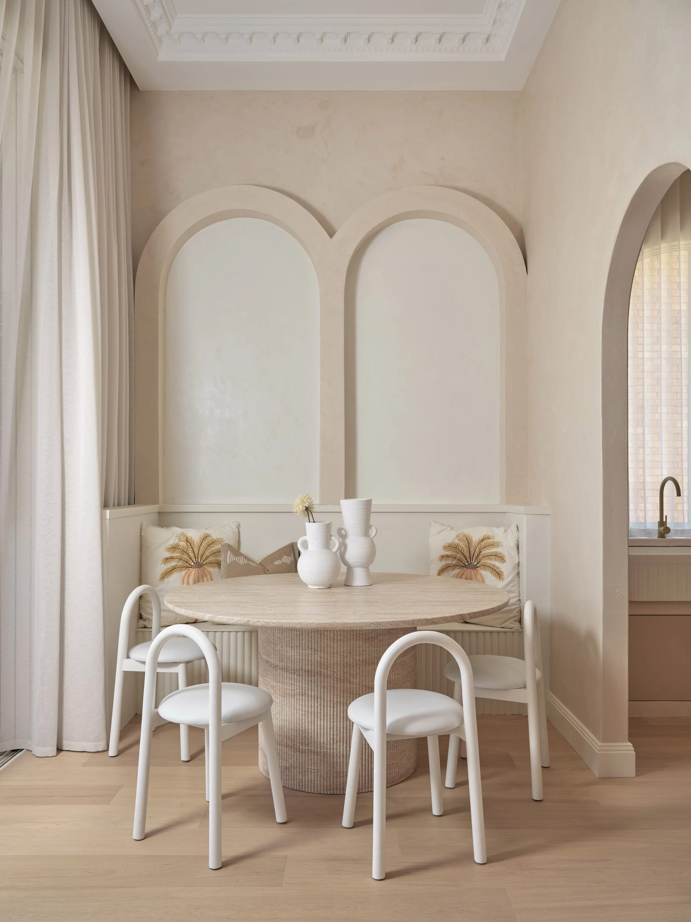 A dining nook with a round table, four white chairs, and beige cushions with palm tree designs, set against a light-colored wall with arched architectural features. Decorative vases with flowers sit on the table.
