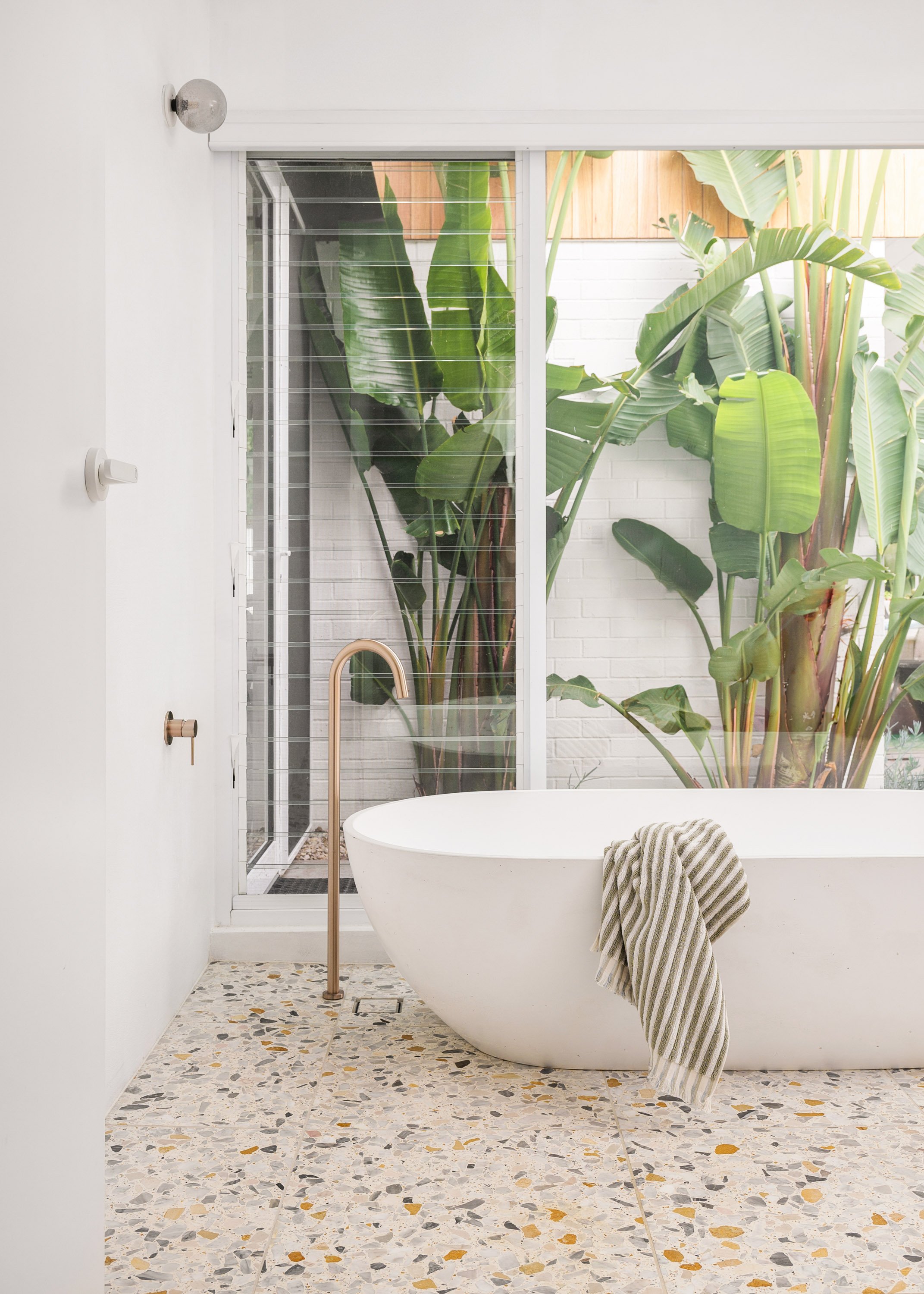 Modern bathroom with a white freestanding bathtub, a striped towel, large green tropical plants outside the window, terrazzo flooring, and a glass shower enclosure.