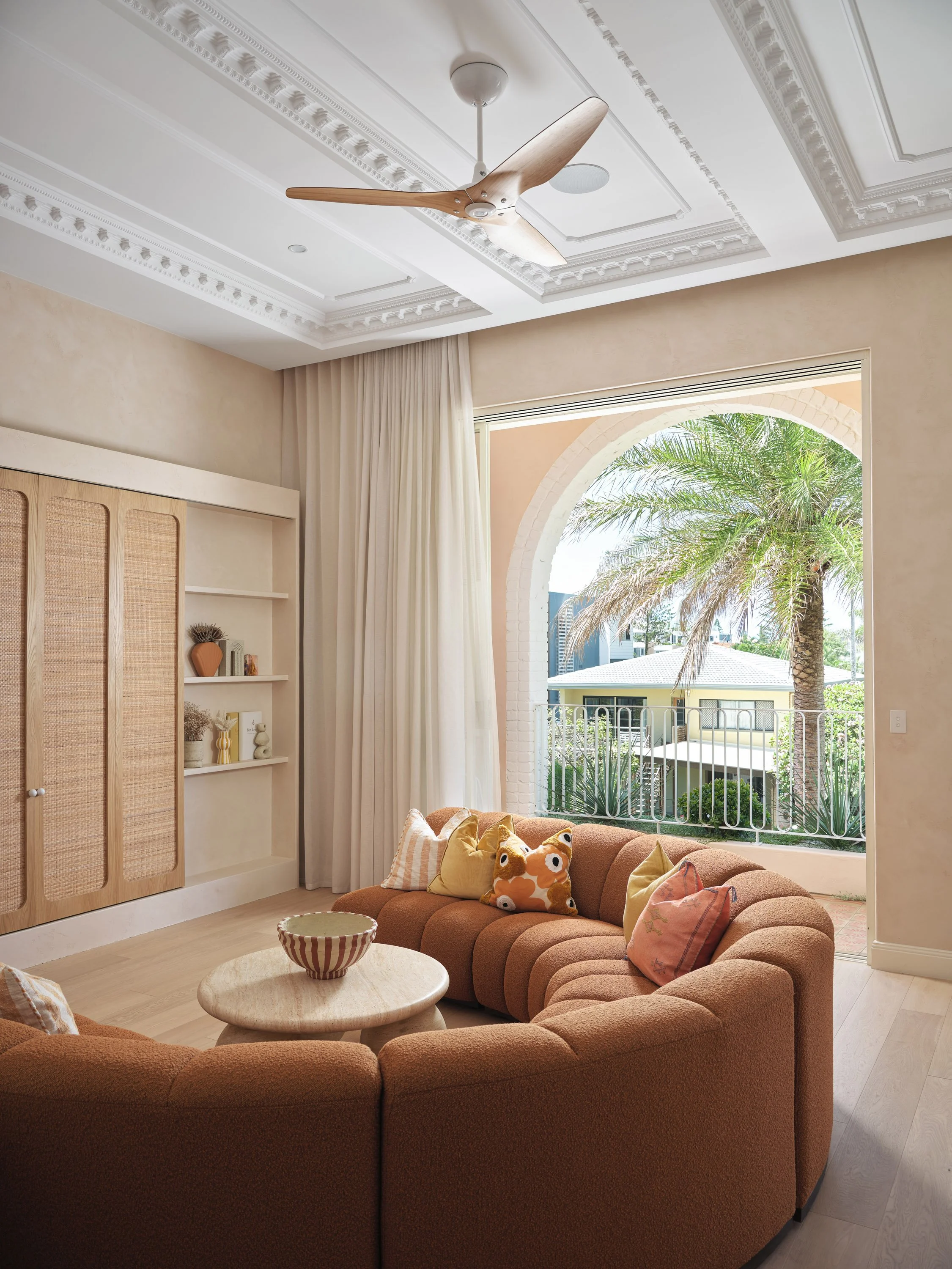 Living room with a brown sectional sofa, decorative pillows, a round wooden coffee table, beige walls, white ceiling with decorative molding, ceiling fan, large open window with white curtains, view of palm tree and neighboring houses outside.